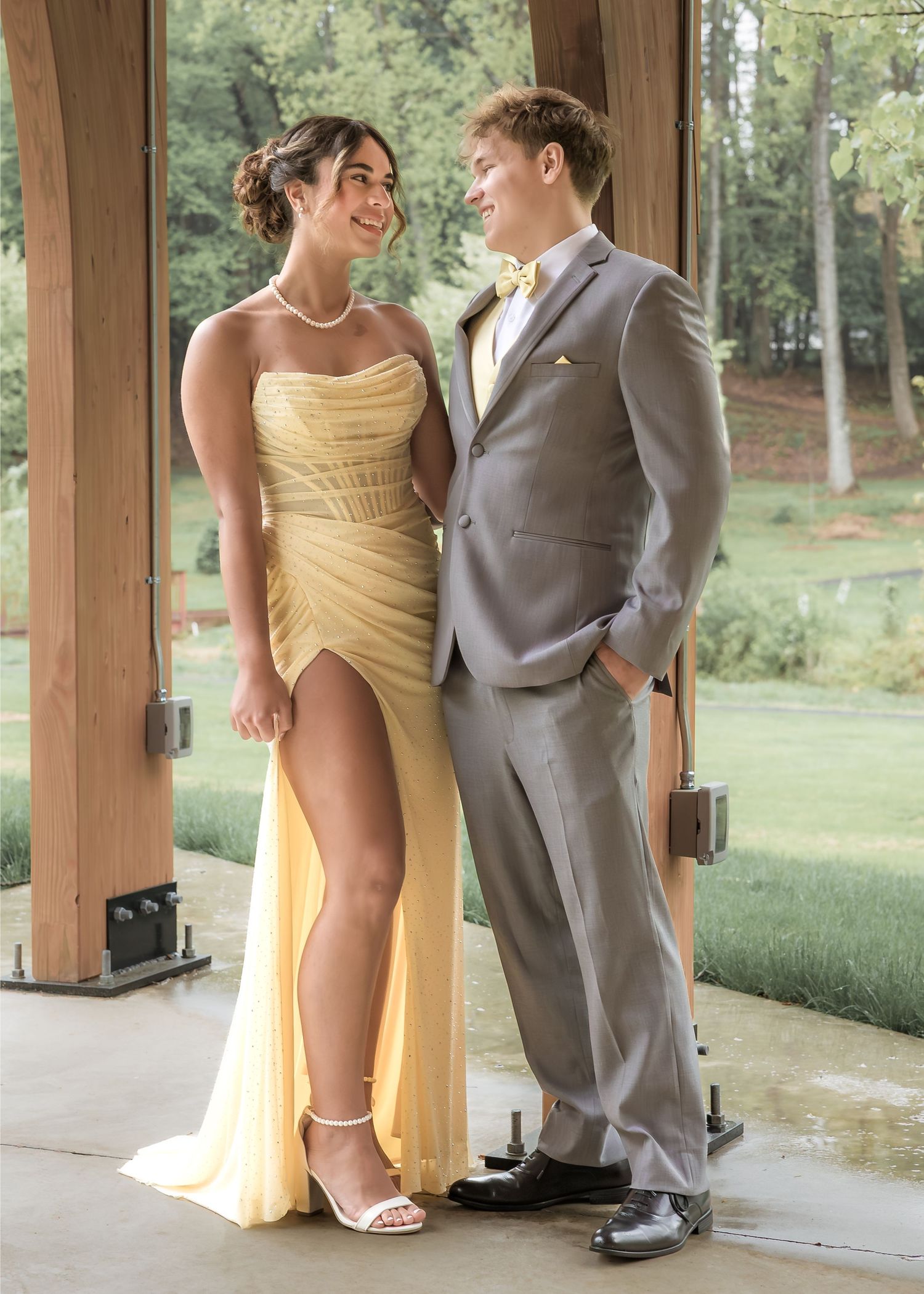 Couple in coordinating yellow prom attire, woman in strapless gown and man in gray suit with bow tie, posing outdoors.