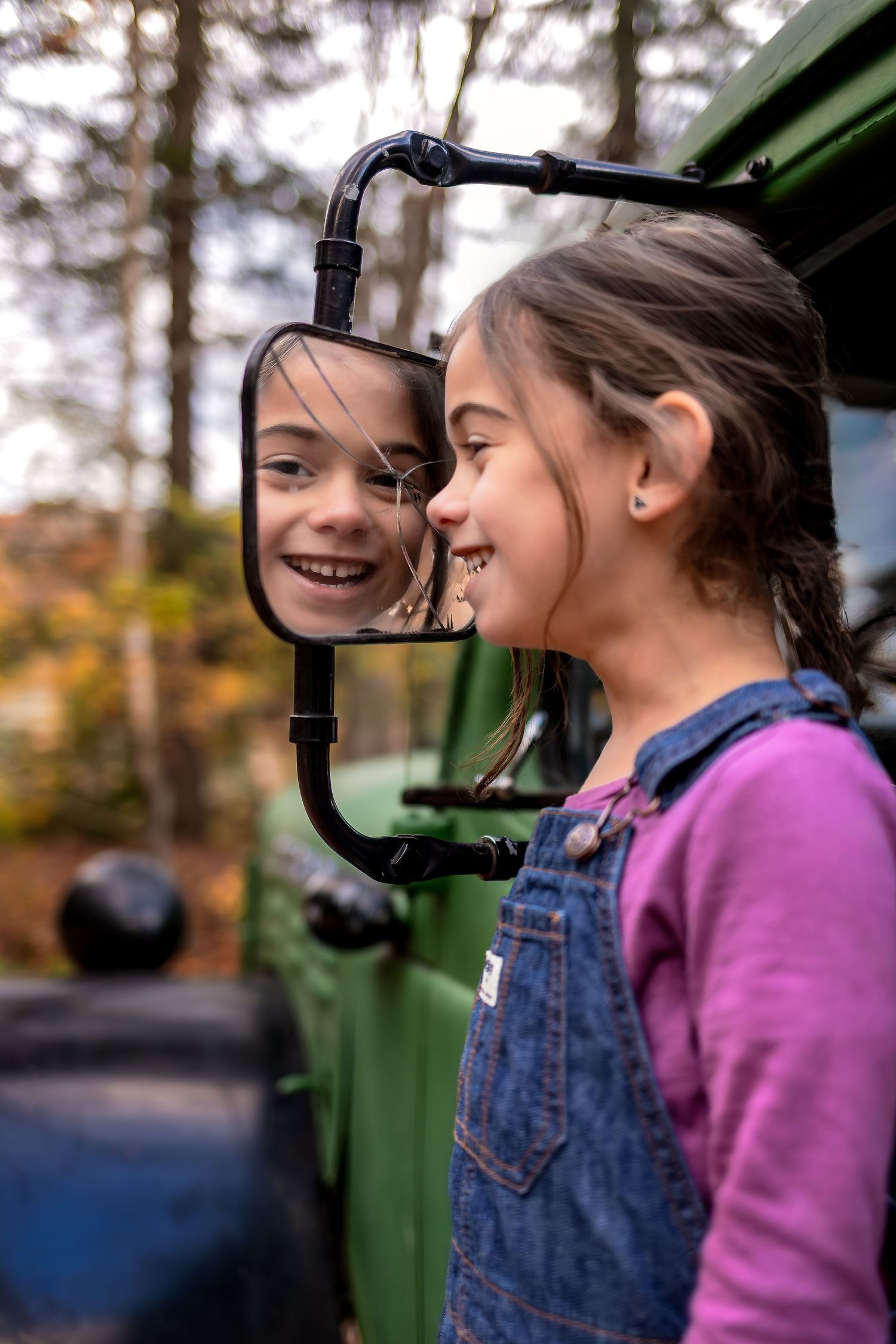 A young girl smiles at her reflection in a vintage green truck mirror during an outdoor autumn photo session.