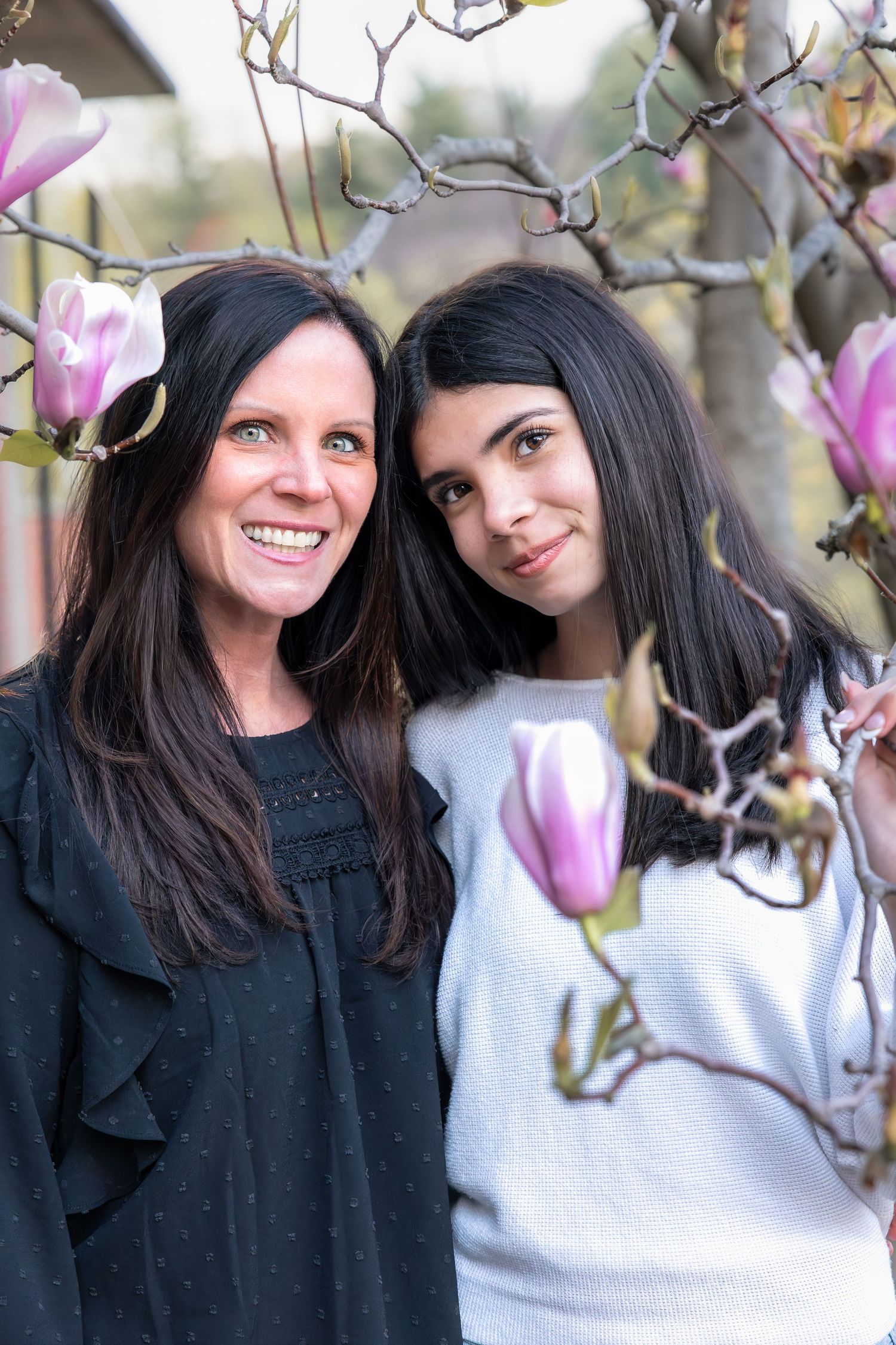 Two women with dark hair smile among pink magnolia blossoms in a spring garden portrait.