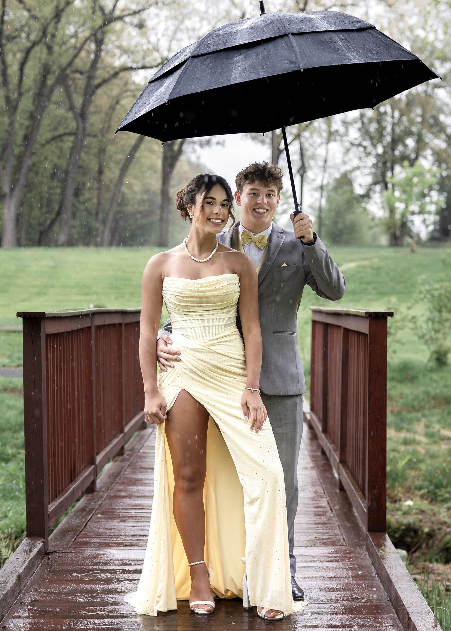 Prom couple in yellow coordinated outfits sharing umbrella on wooden bridge in the rain.