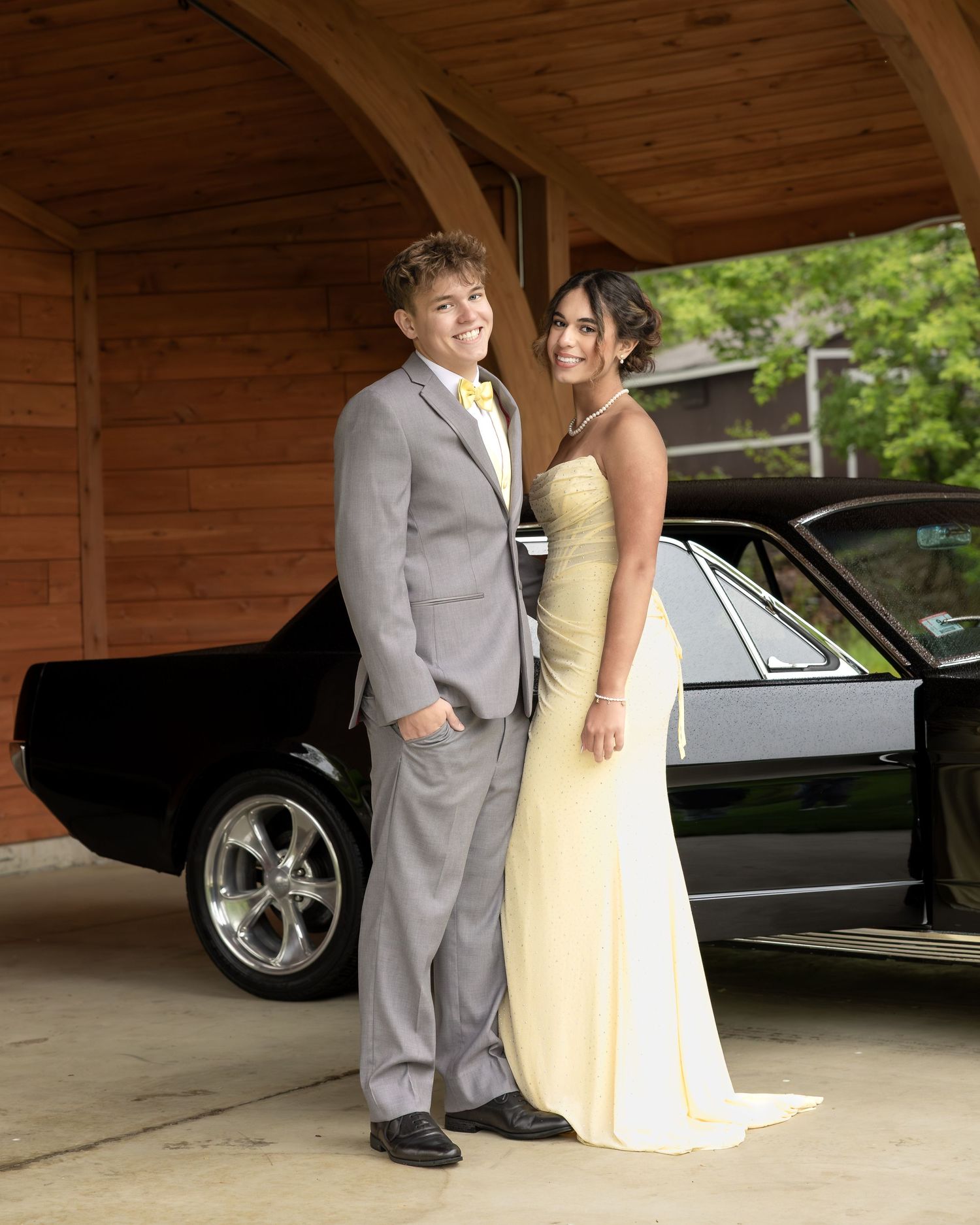 A young couple in prom attire, gray suit and yellow gown, posing beside a classic black car under a wooden structure.