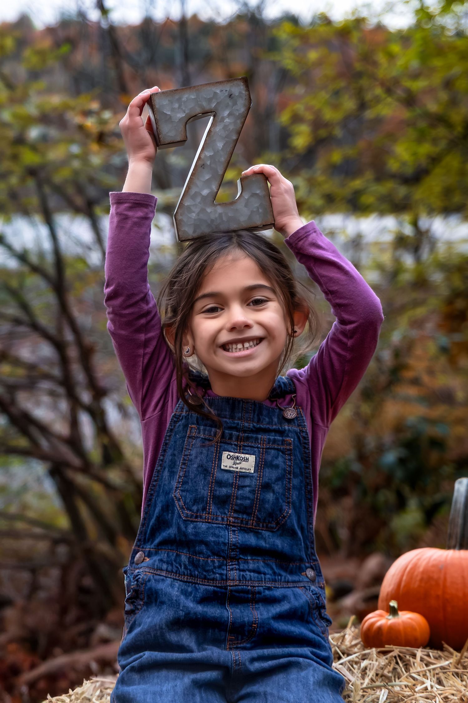 Smiling girl in denim overalls holds metal letter Z above her head while sitting on hay bales with pumpkins in fall.