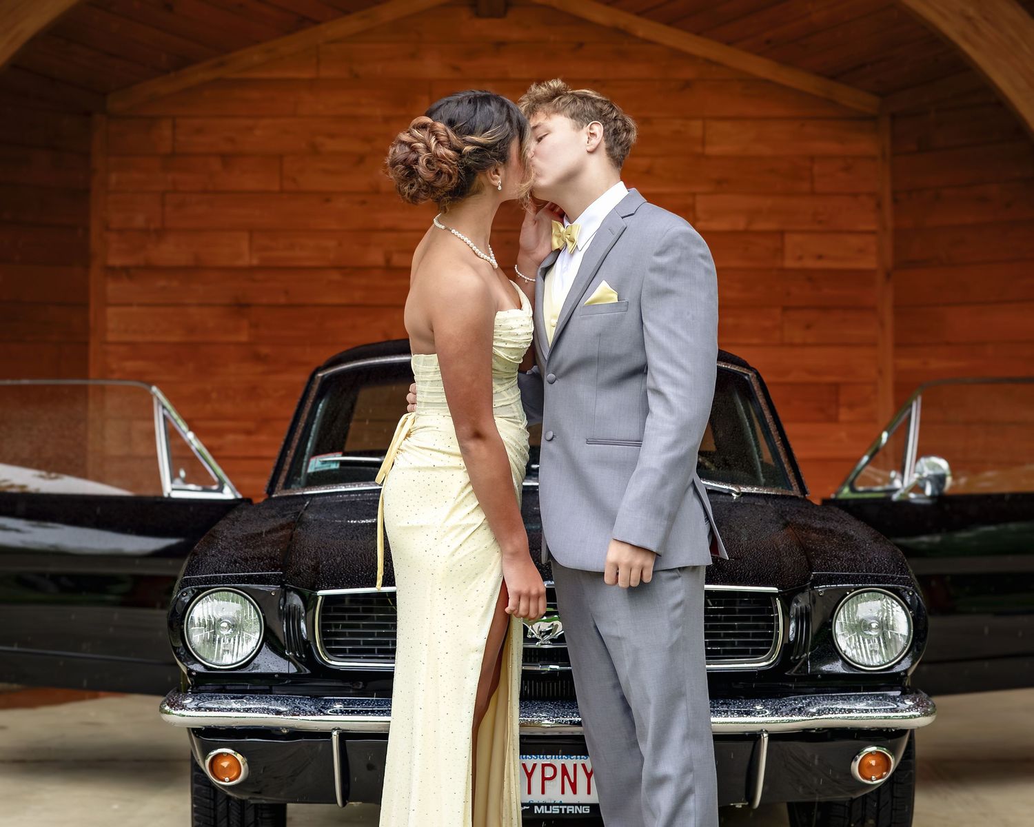 Couple in formal wear sharing a kiss in front of a classic black Ford Mustang near a wooden garage.