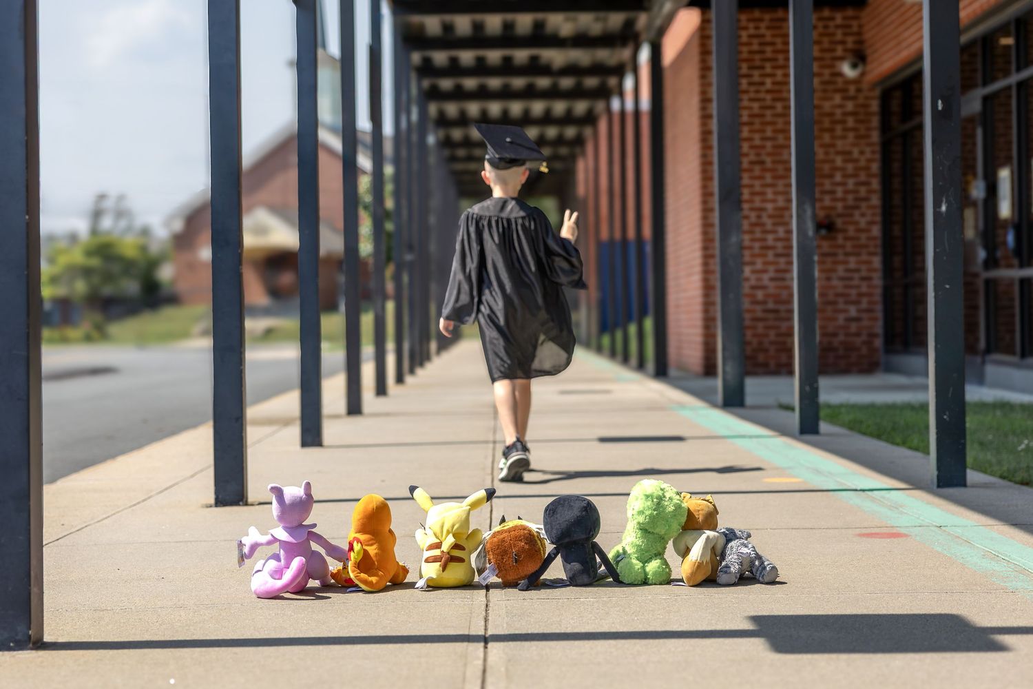 Child in graduation gown walks past a row of colorful stuffed Pokemon toys lined up on a school sidewalk.