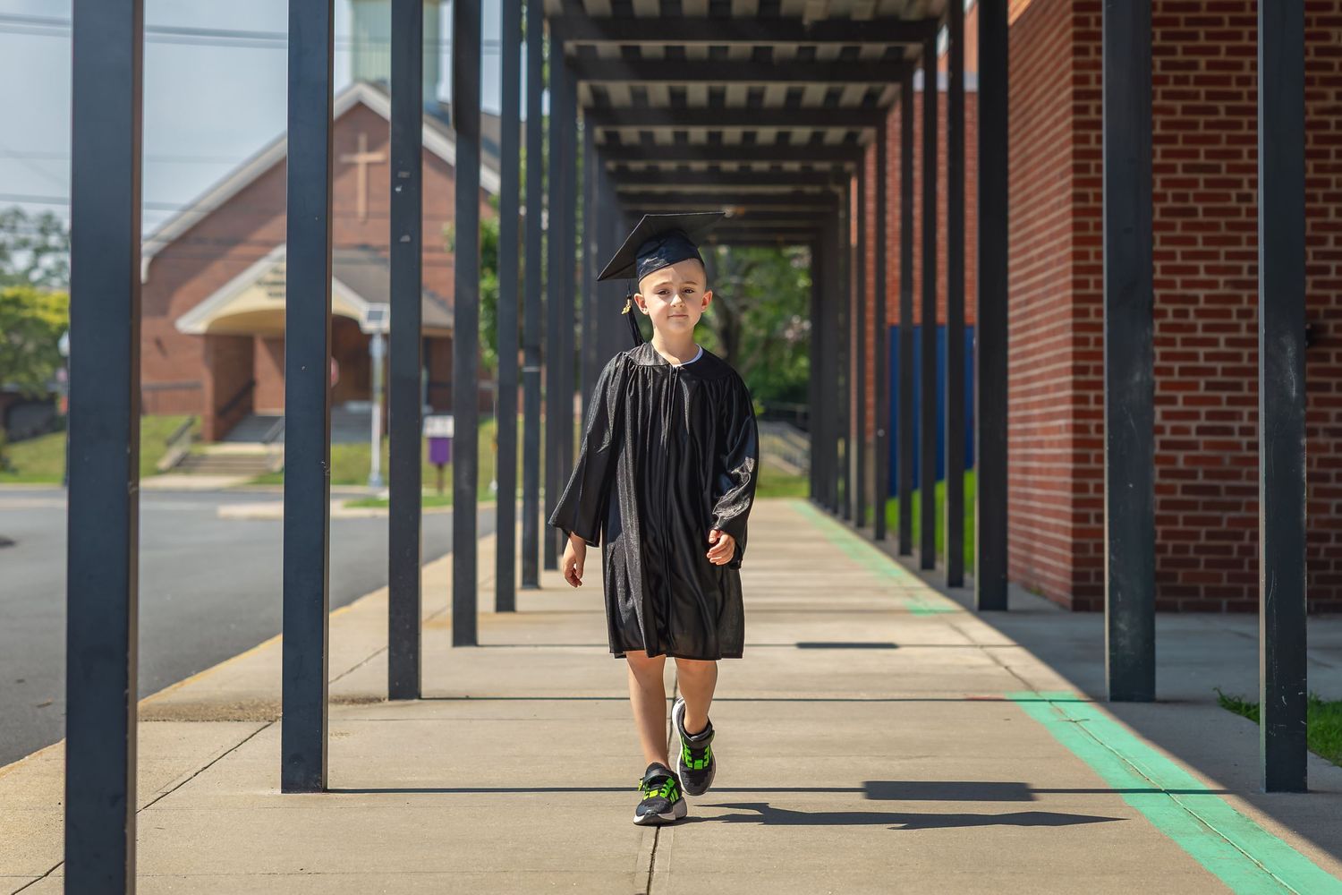 Young child in black graduation cap and gown walks confidently along a brick school building colonnade.