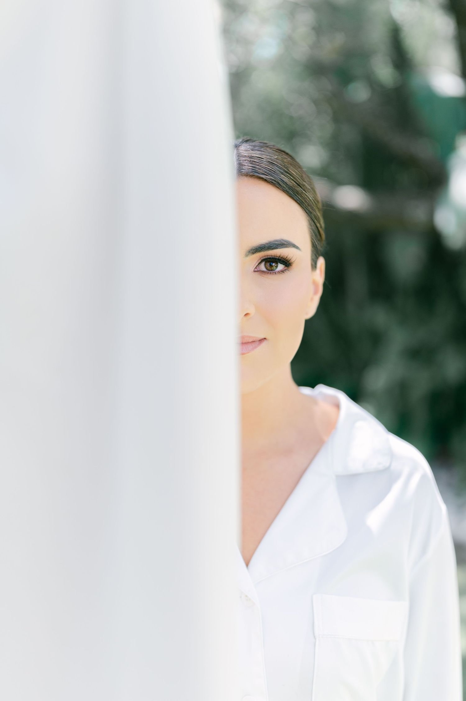 Bride in white robe peeking from behind white curtain with lush green trees in soft background.