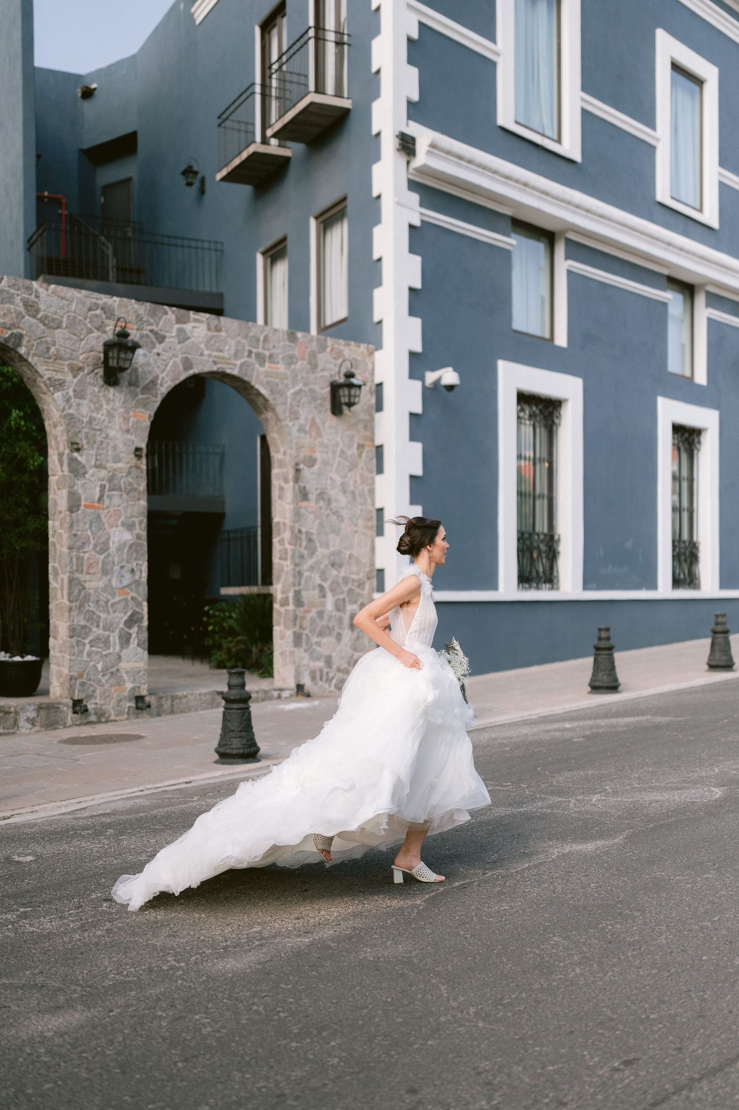 Bride in flowing white wedding gown crossing a street beside a blue stone building.