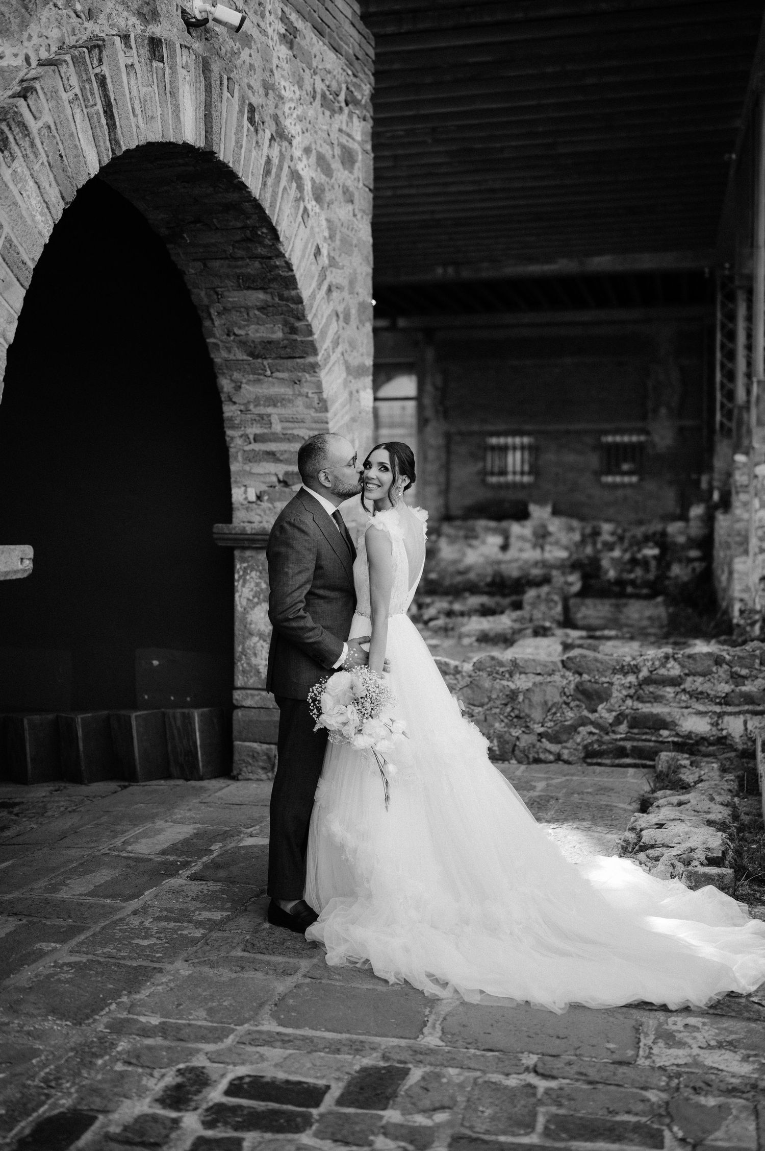 Black and white photo of groom kissing bride near a stone arch at a rustic ruins wedding venue.
