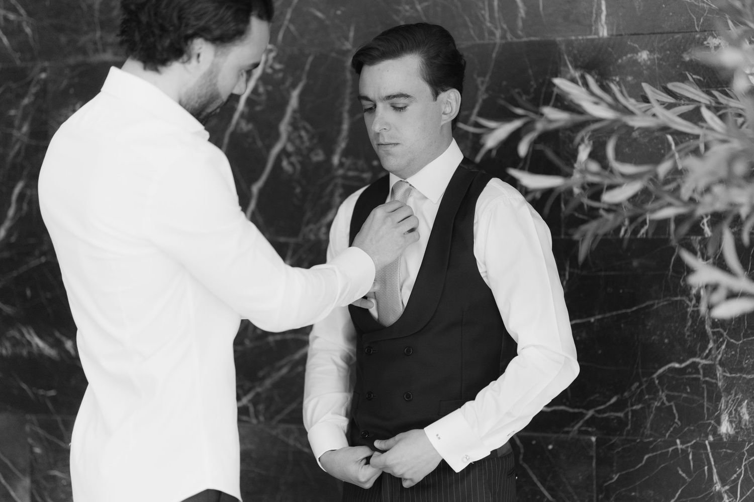 Black and white photo of a man adjusting another mans tie, both dressed in formal wedding attire.
