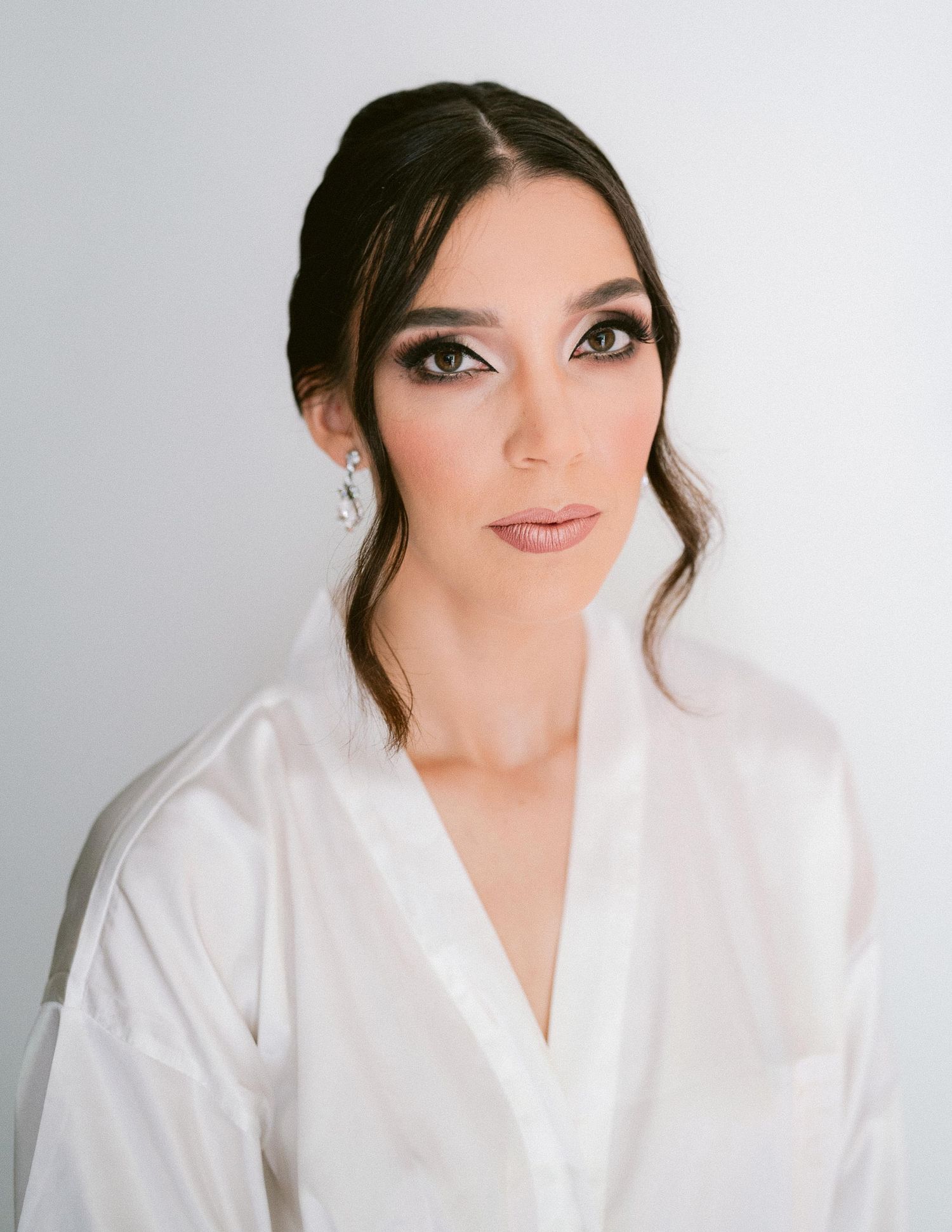 Stunning bride with dark updo, smoky eye makeup, and crystal earrings wearing a white silk bridal robe.
