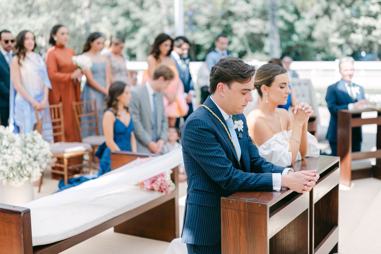 Bride and groom kneeling in prayer at outdoor wedding ceremony with guests standing behind.