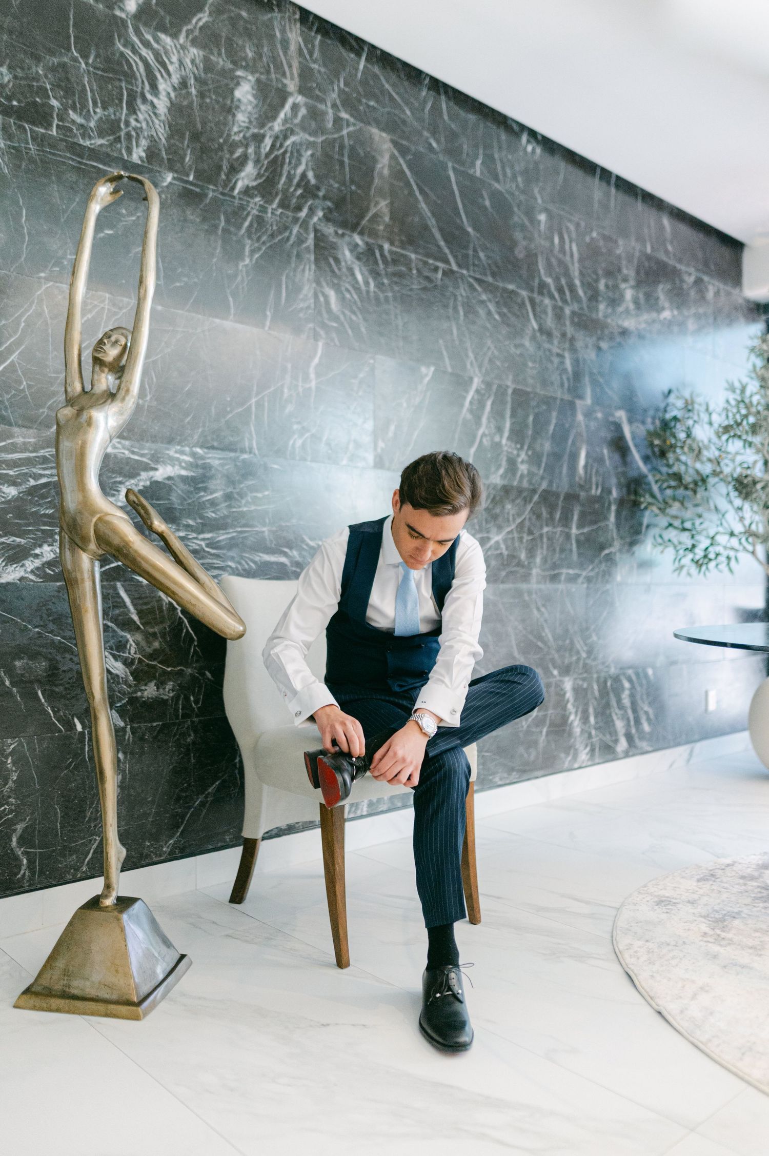 Groom in navy suit and vest fastening his shoe beside a bronze ballerina statue on marble floor.
