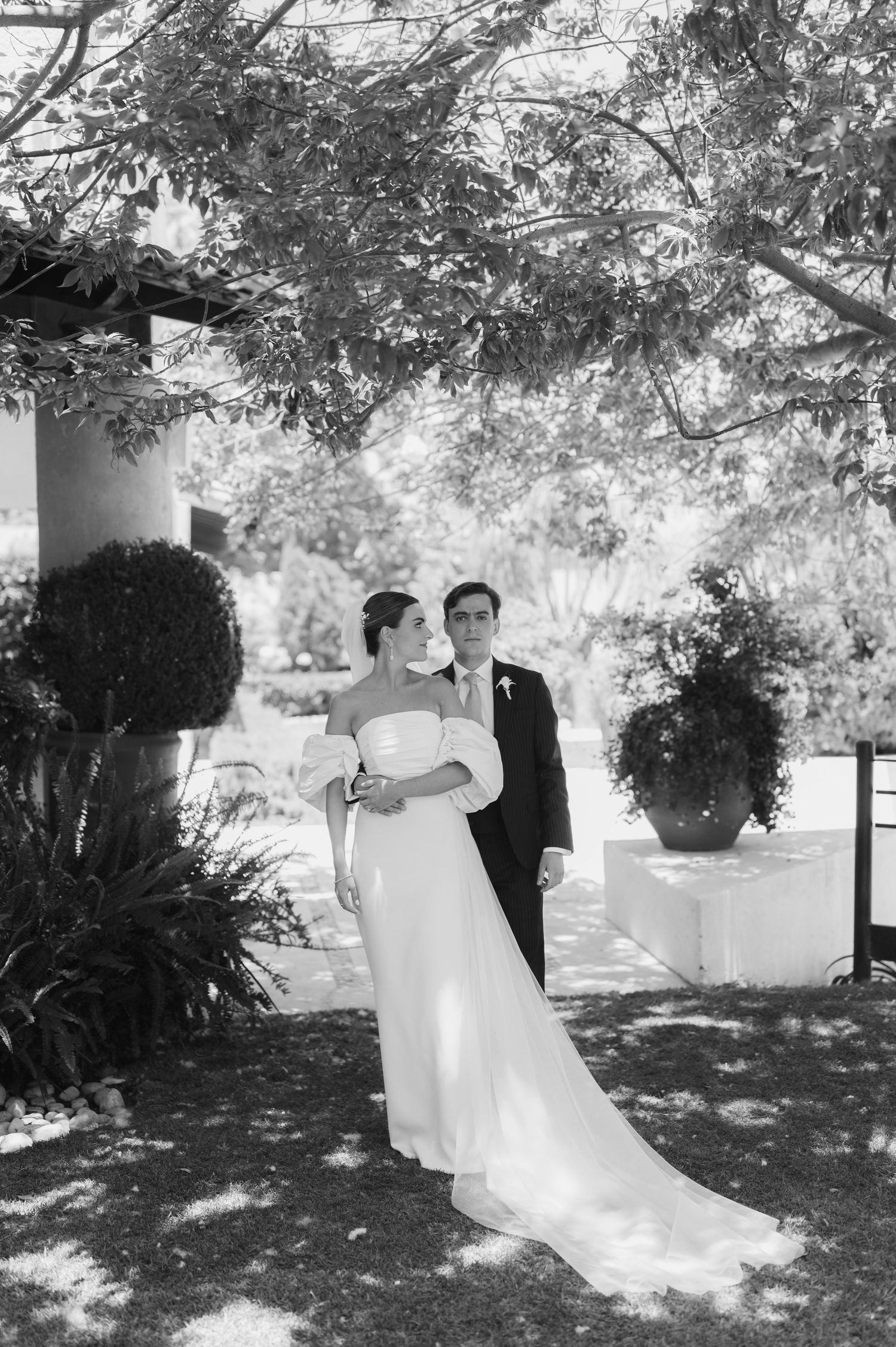 Black and white photo of bride in off-shoulder gown and groom in suit posing under trees at outdoor wedding venue.