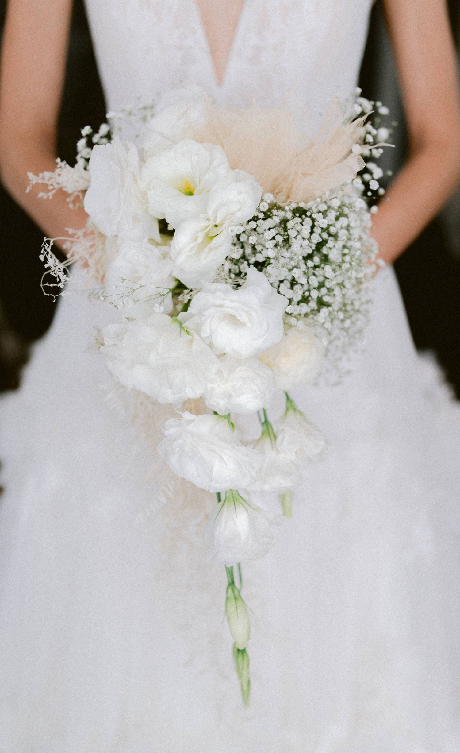 Cascading bridal bouquet of white lisianthus and baby's breath held by bride in lace gown.