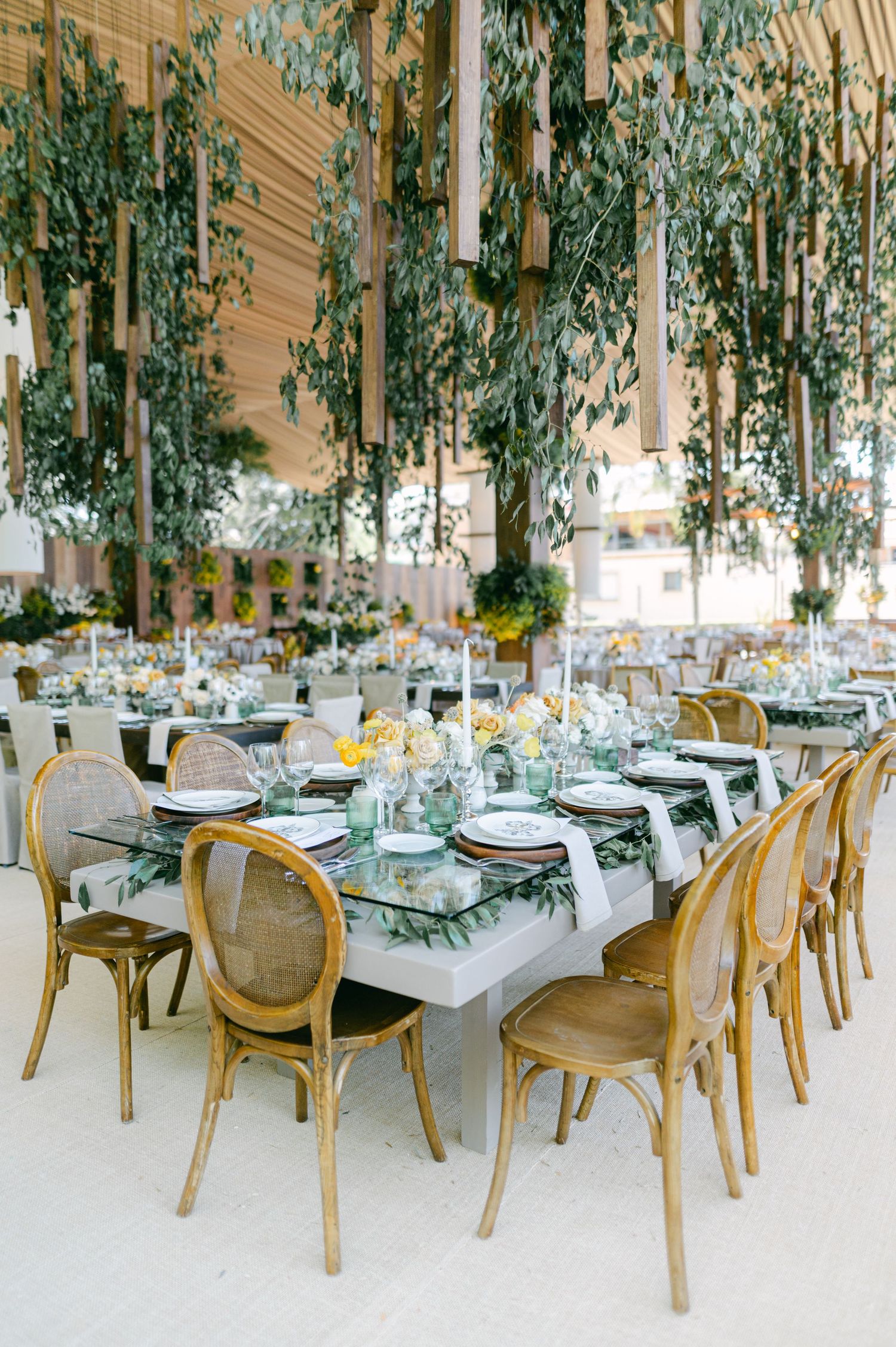 Long wedding reception table with greenery runner and rattan chairs under hanging foliage.