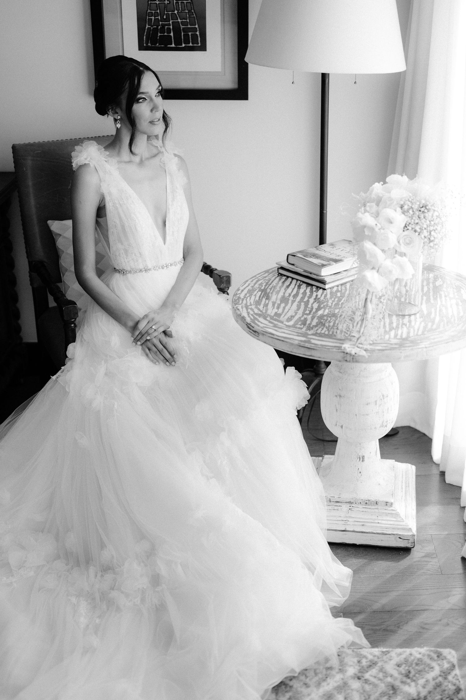 Black and white photo of seated bride in voluminous tulle gown beside ornate side table.