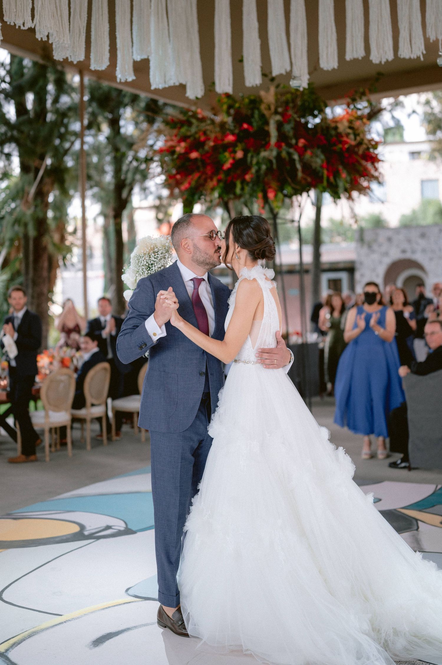 Bride and groom share a romantic first dance kiss in a white wedding gown and navy suit at an outdoor reception.