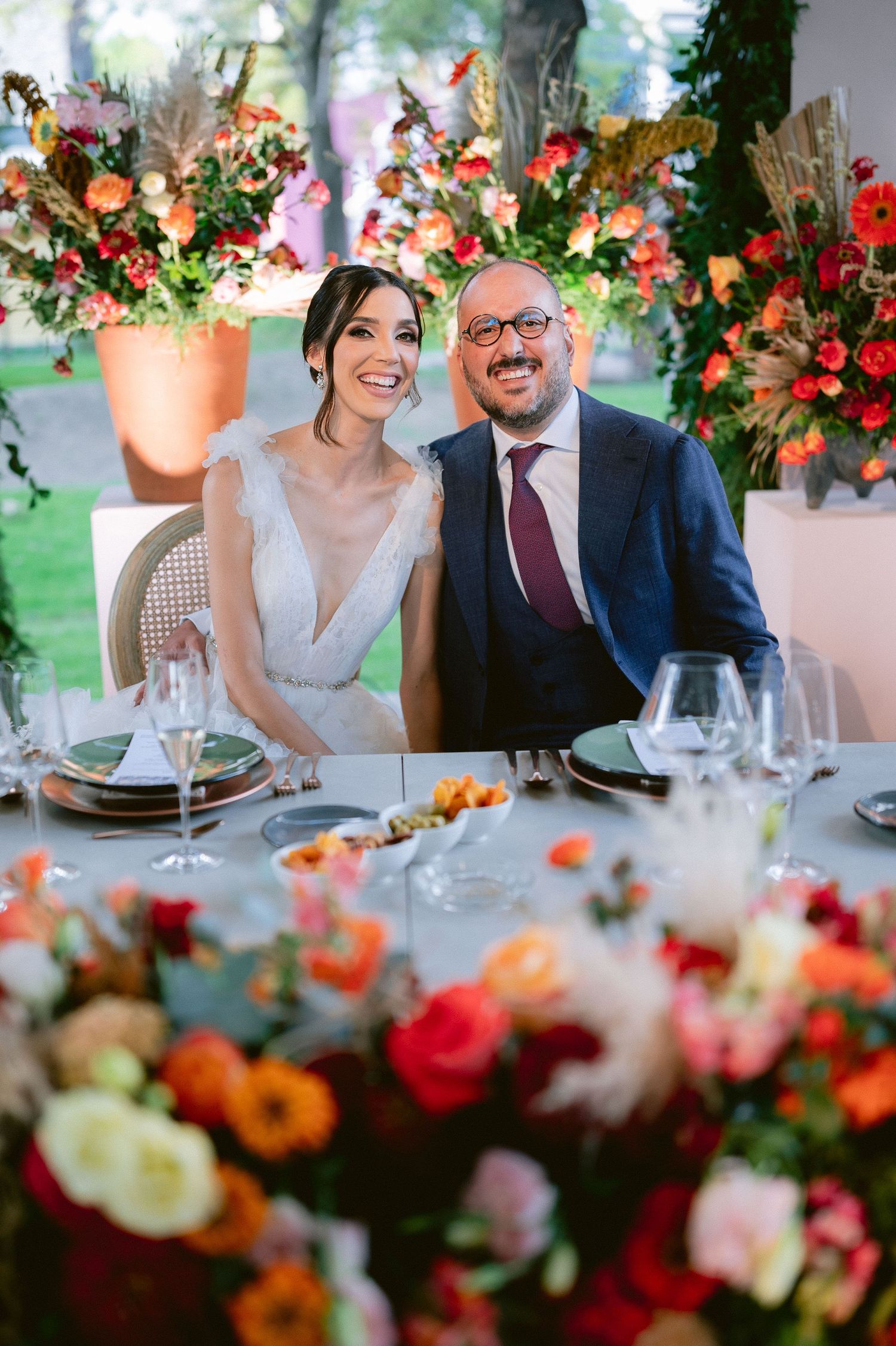 Happy bride and groom pose together at a sweetheart table surrounded by lush orange and red floral centerpieces.