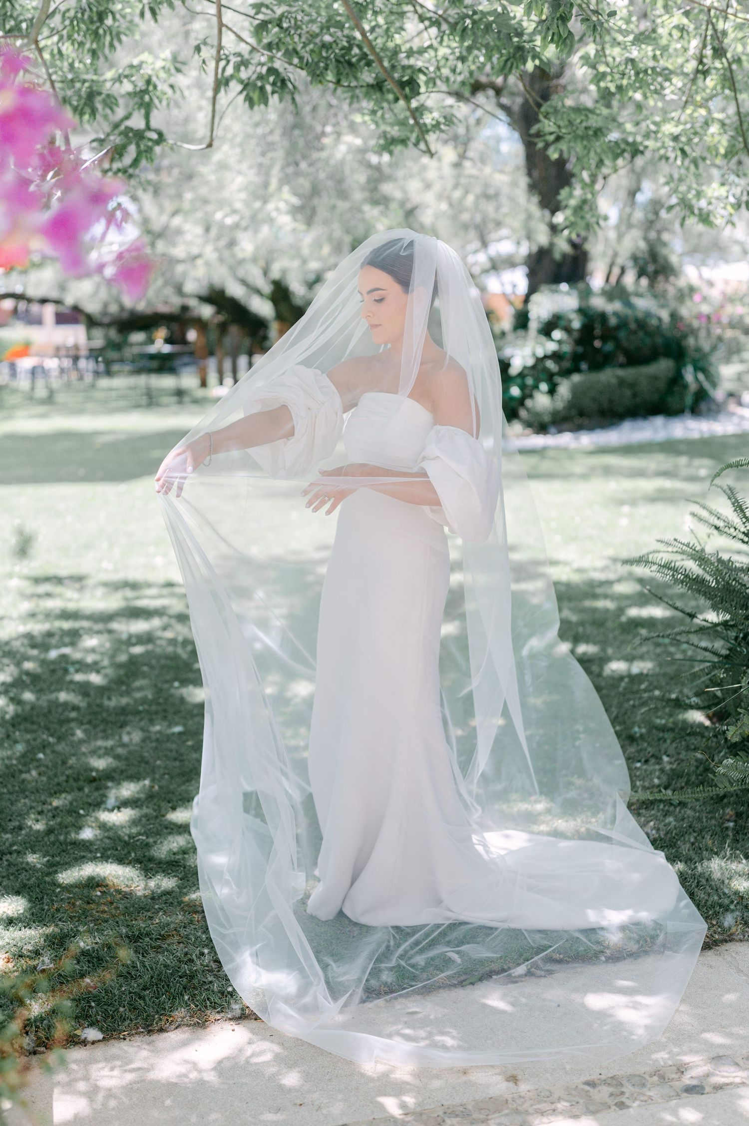 Bride in elegant white wedding gown holding flowing veil in lush garden setting.