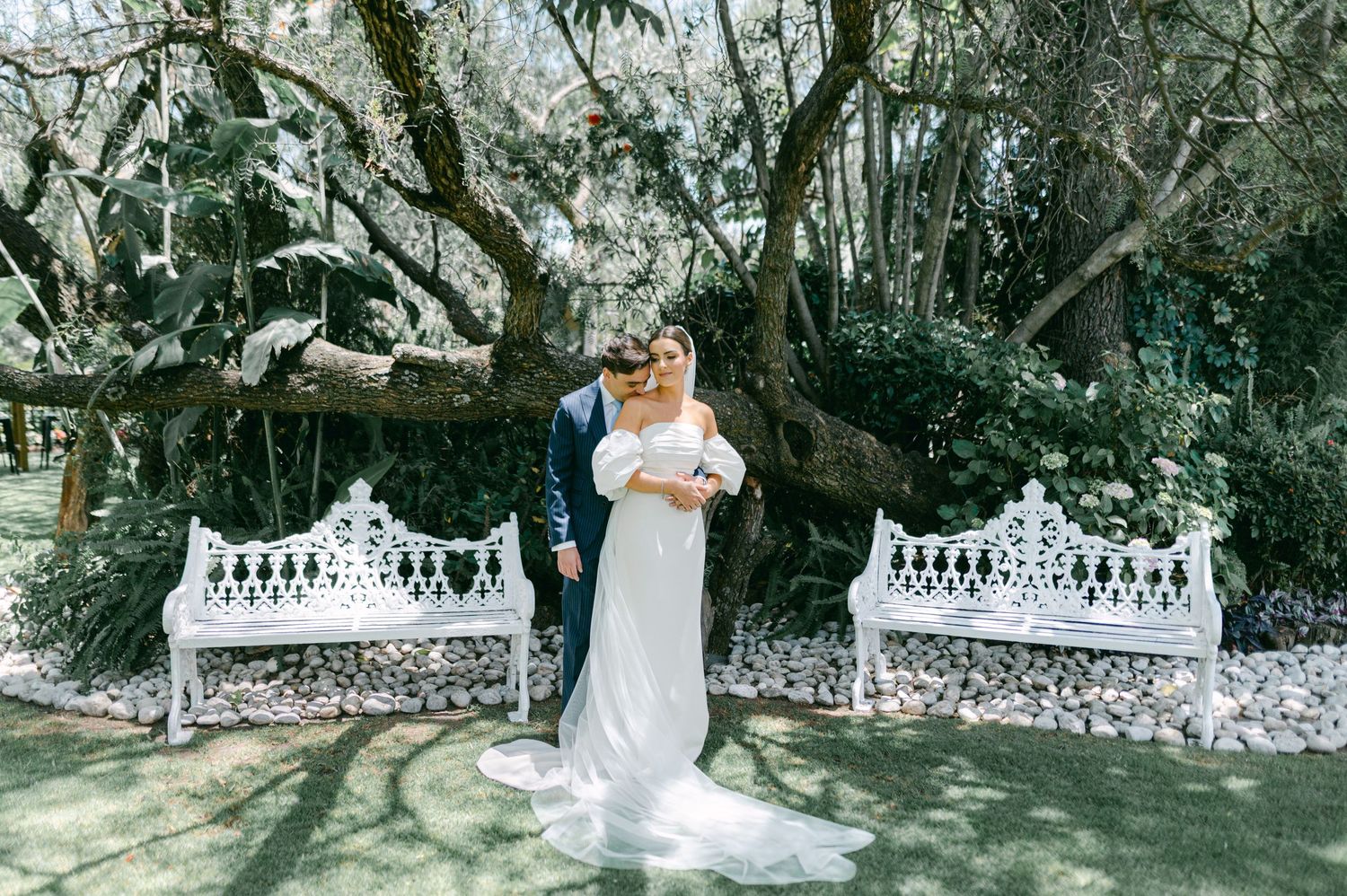 Bride and groom posing before large oak tree with white ornate benches in lush garden wedding setting.