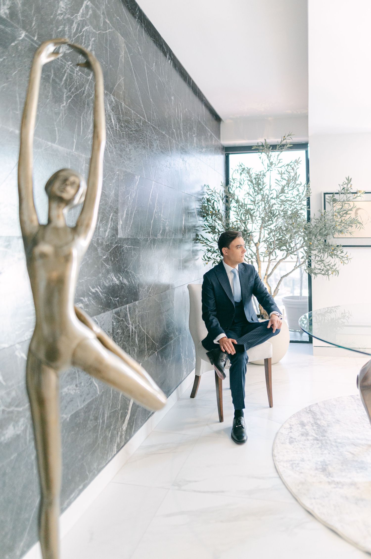 Groom in navy suit seated beside a bronze ballerina sculpture in a modern marble interior.