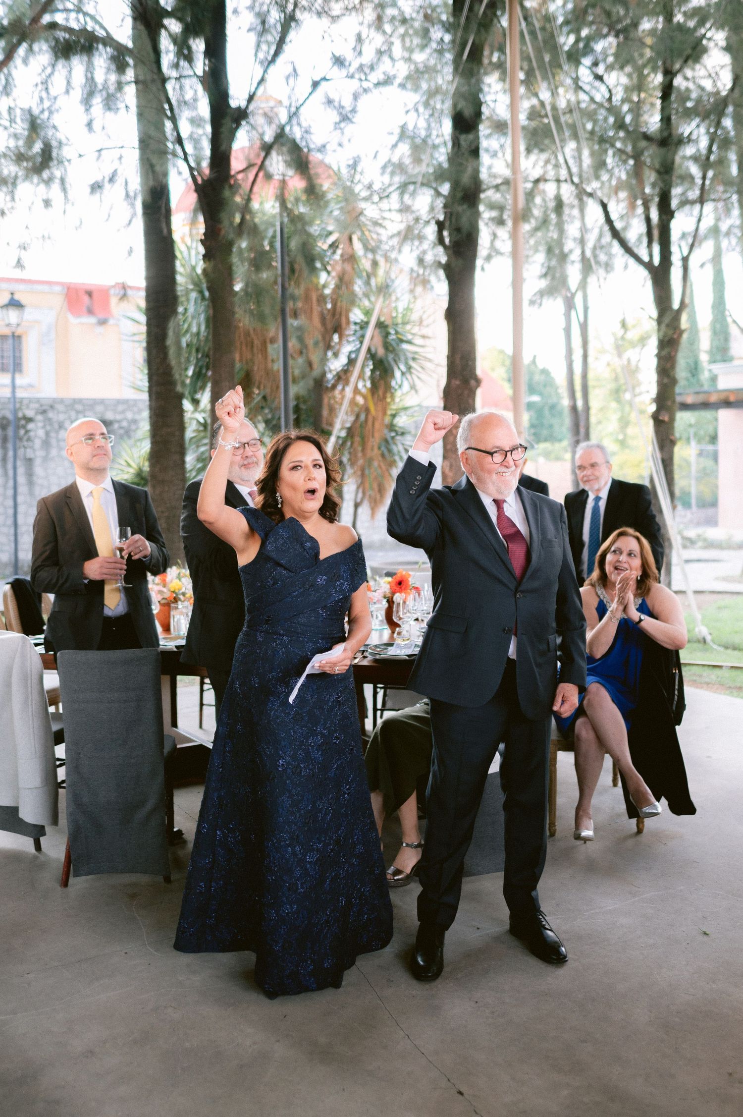 Elderly couple celebrates joyfully with raised fists at an outdoor wedding reception surrounded by guests.