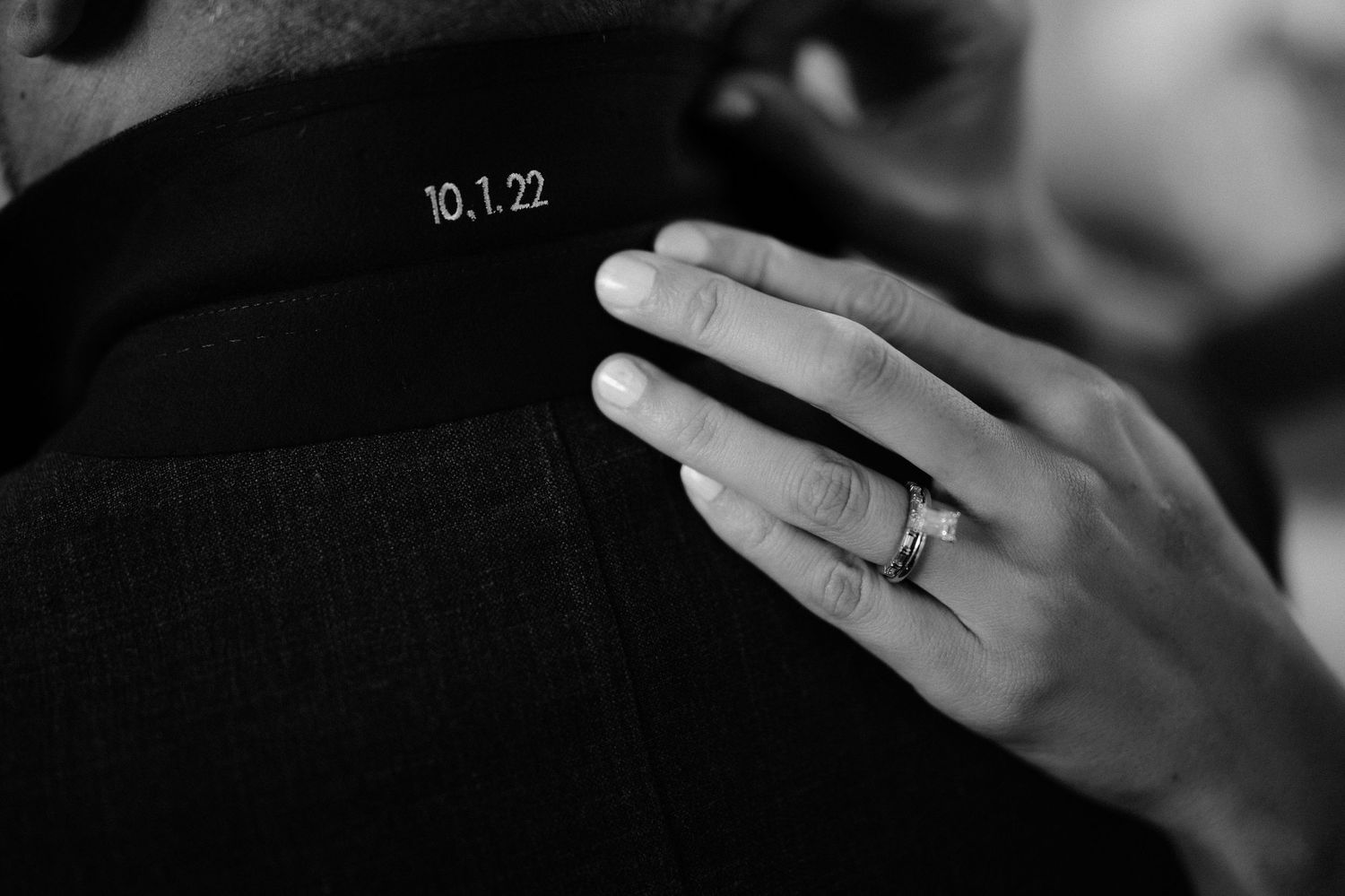 Close-up black and white photo of a bride's diamond ring resting on groom's jacket labeled 10.1.22.