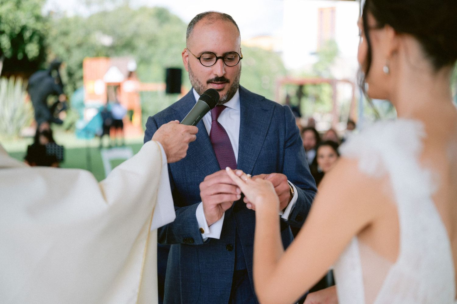 Groom places wedding ring on brides finger during outdoor ceremony while officiant holds microphone.