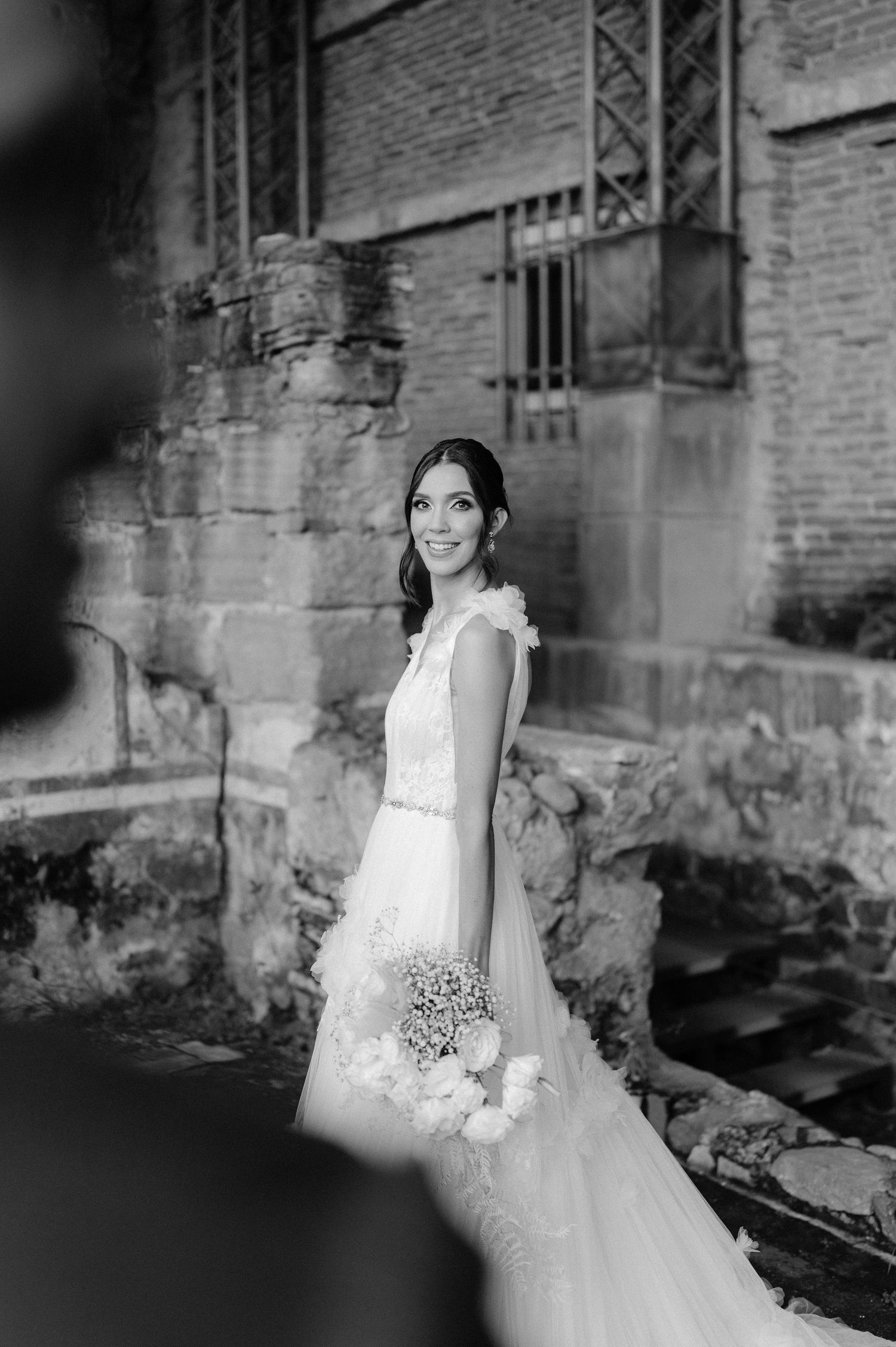 Black and white portrait of smiling bride in lace wedding gown holding bouquet at brick ruins venue.