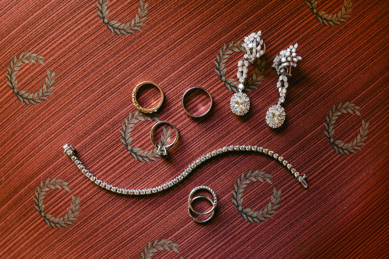 Elegant wedding jewelry flat lay featuring diamond earrings, tennis bracelet, and gold rings on red patterned surface.