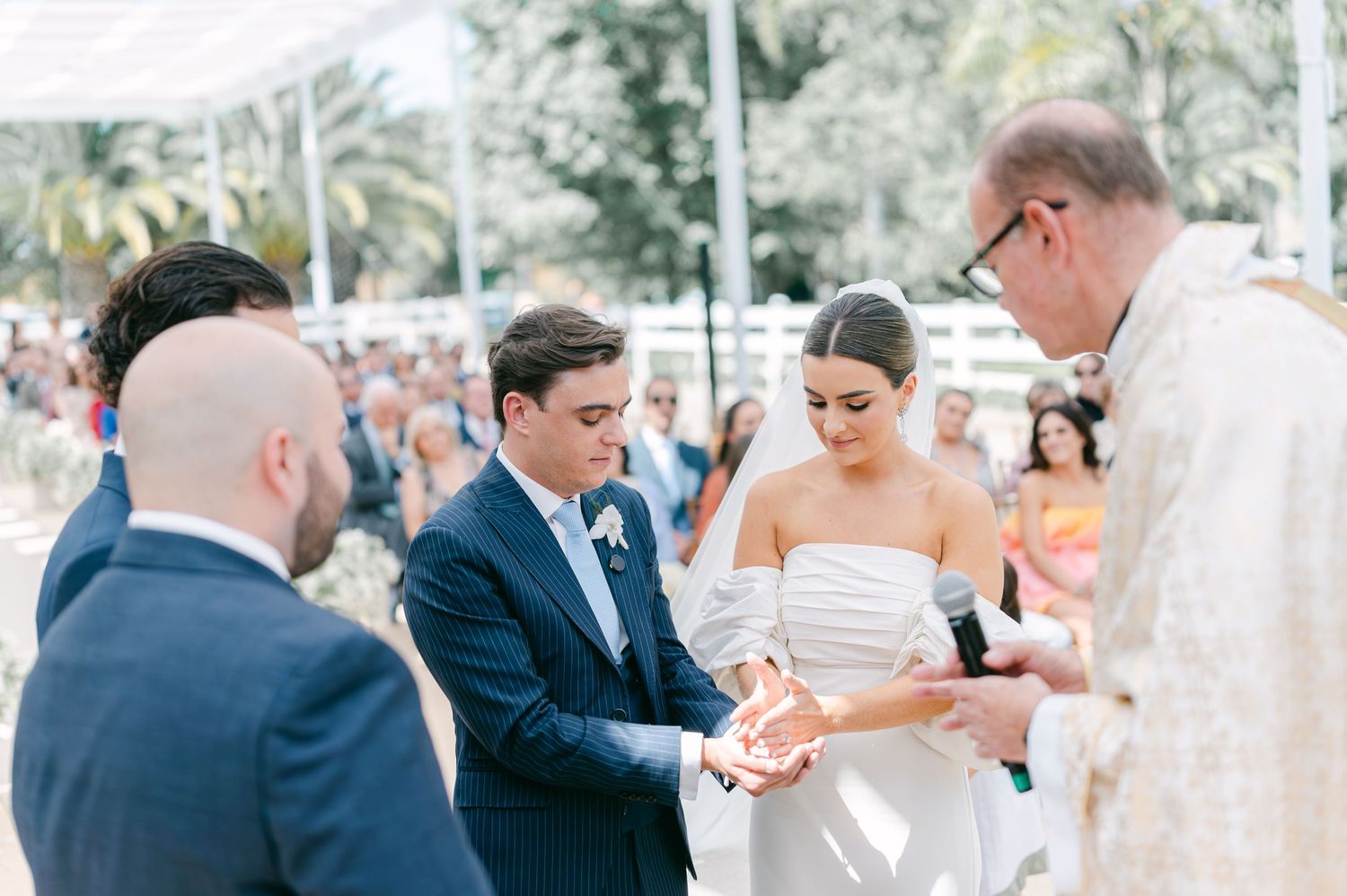 Priest blessing bride and groom during outdoor wedding ring exchange ceremony.