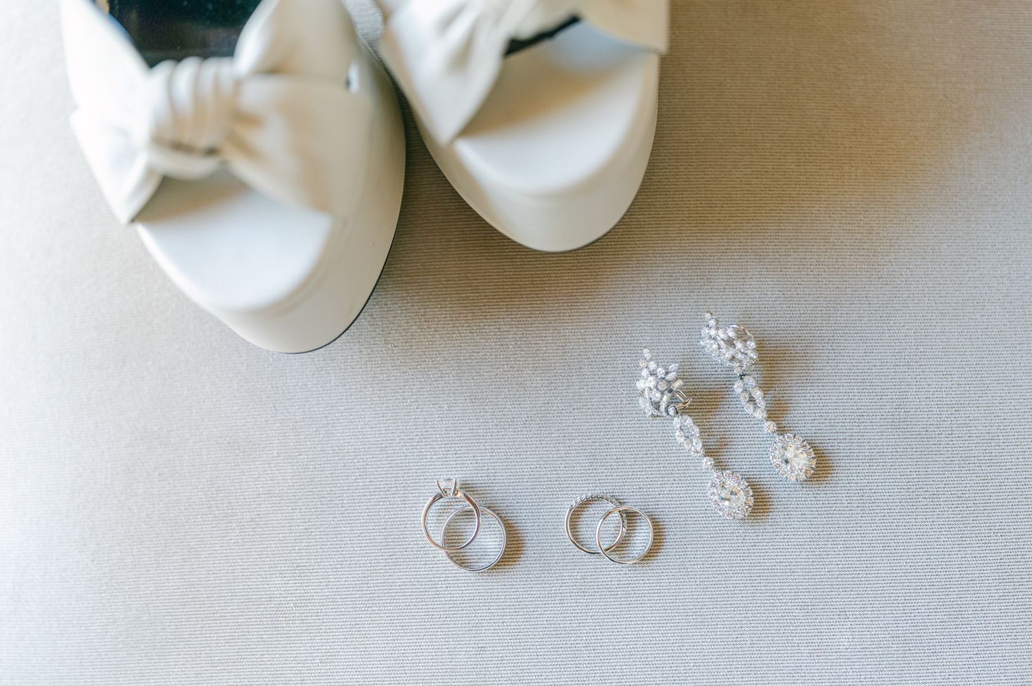 White bow bridal heels displayed alongside diamond drop earrings and wedding bands on neutral surface.
