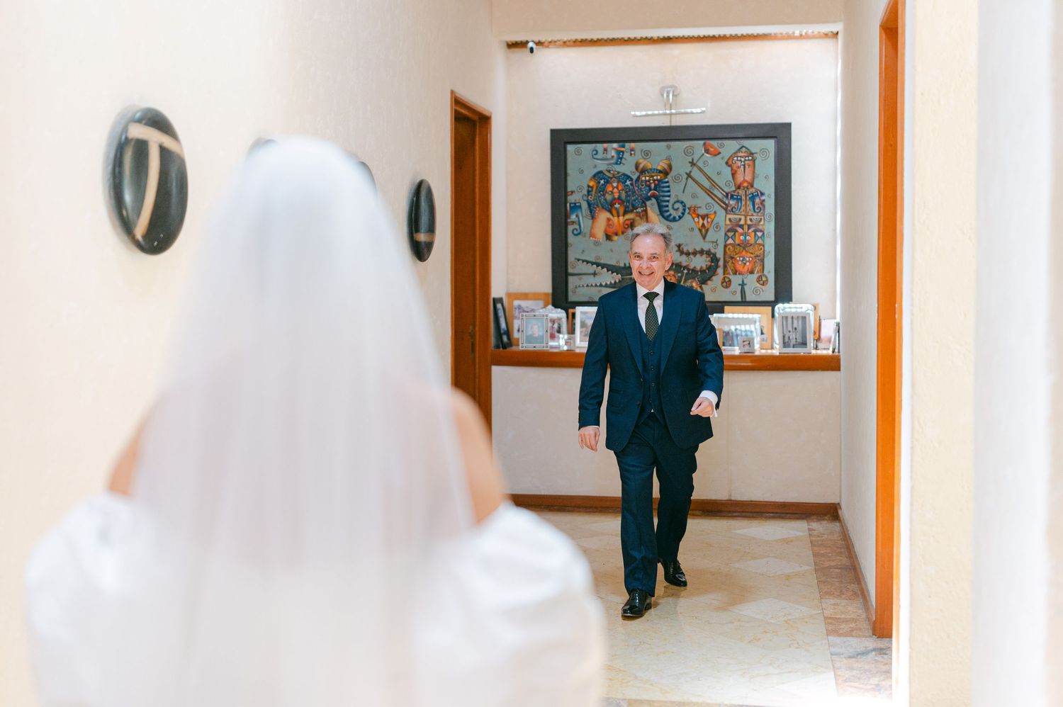 Father walking down hallway toward bride in white wedding dress and veil before ceremony.