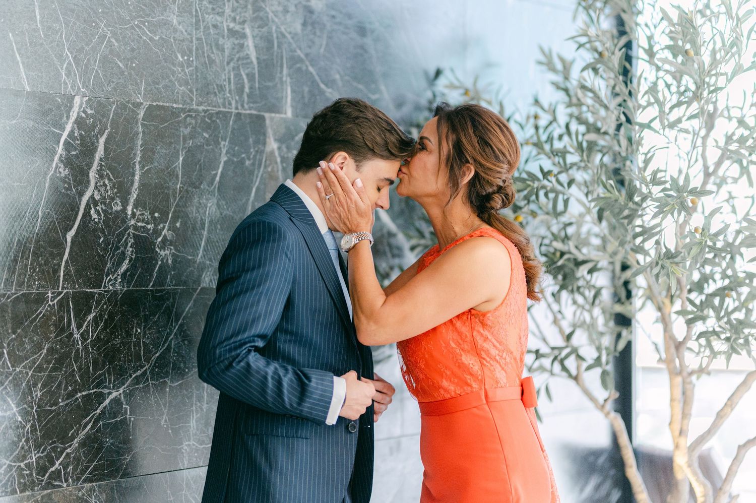 Couple sharing an intimate forehead touch moment, woman in orange dress beside marble wall.