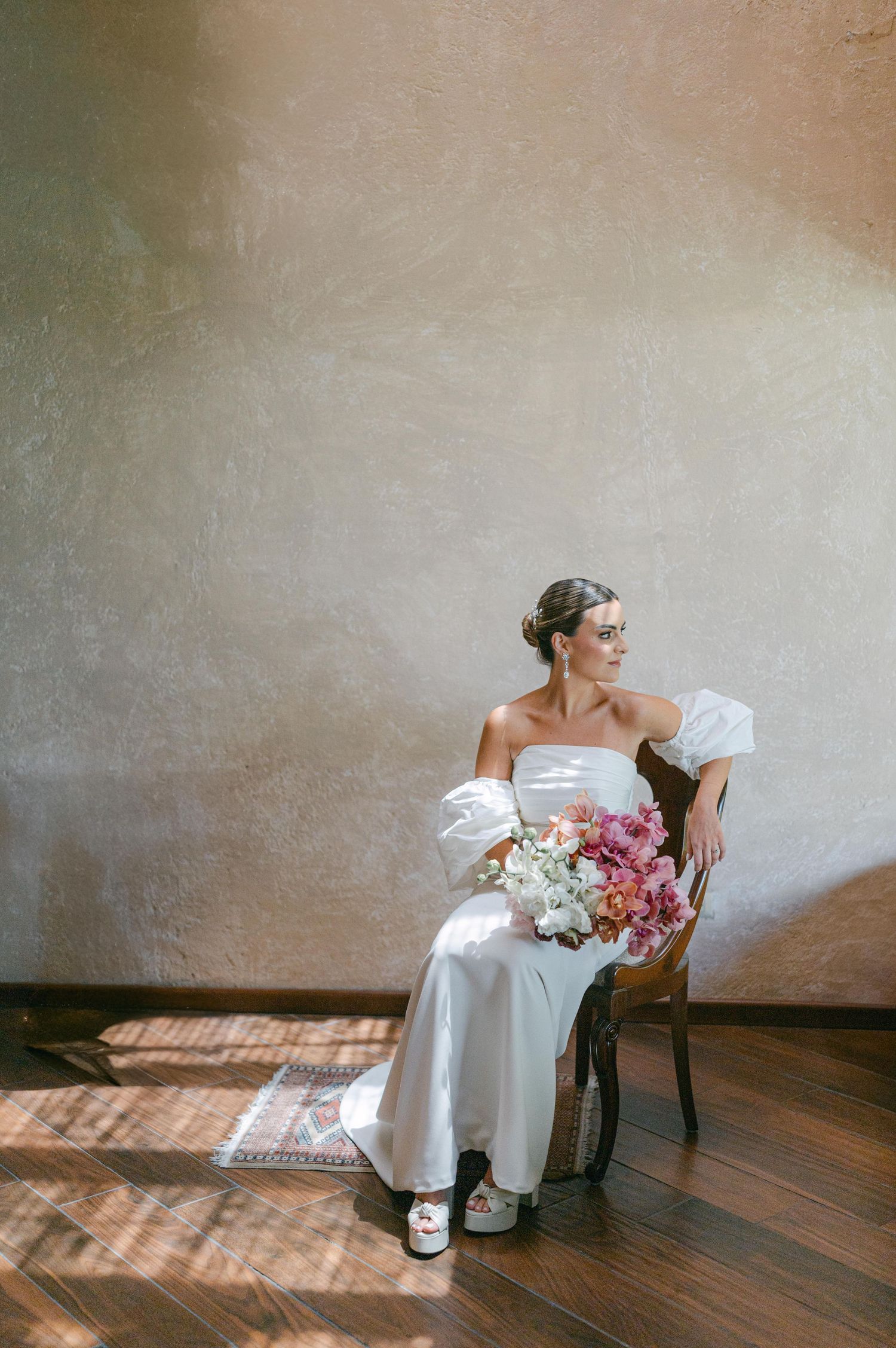 Bride in off-shoulder white satin gown with puff sleeves sits in chair holding pink and white floral bouquet.