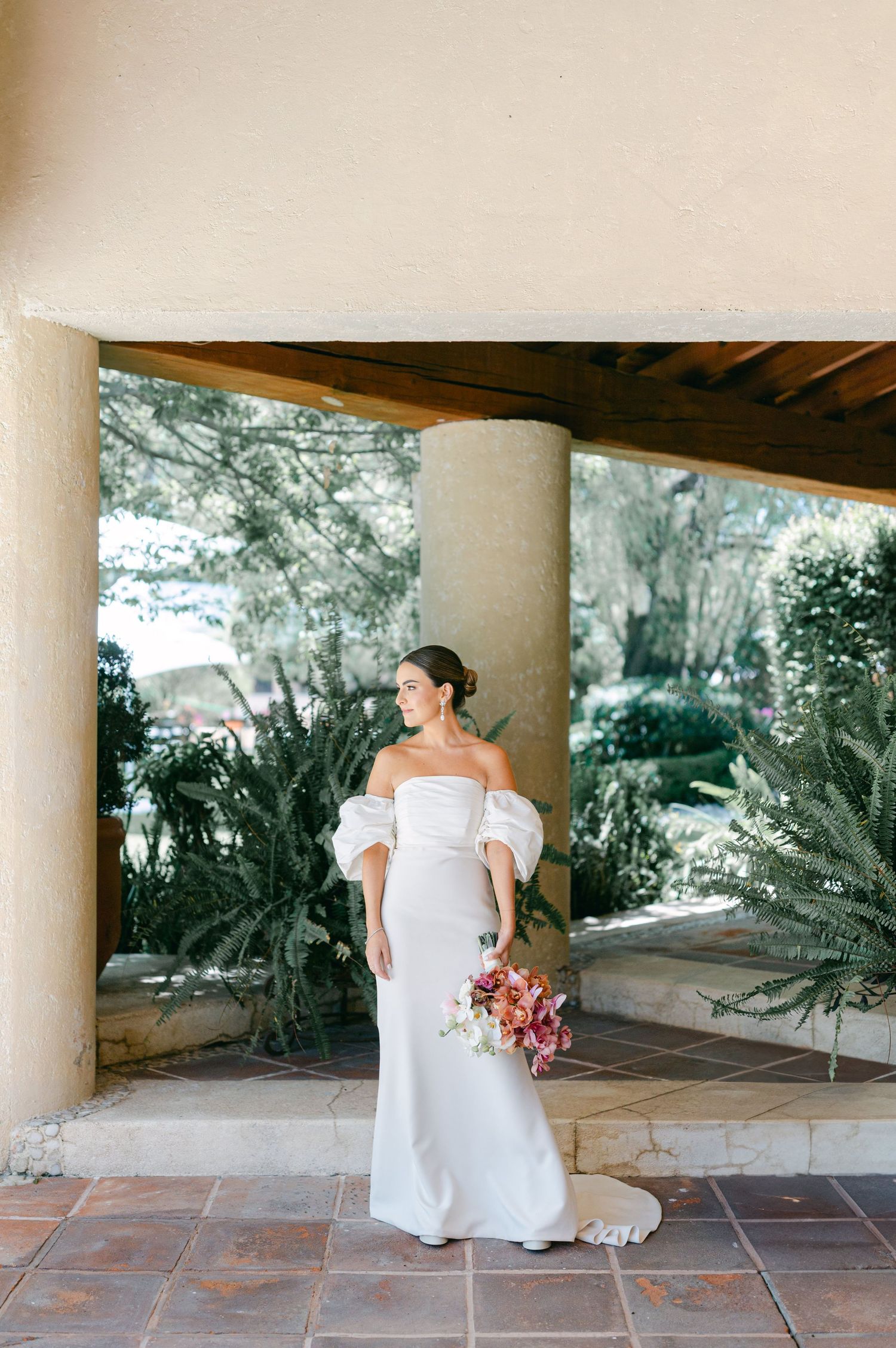 Bride in elegant off-shoulder white gown with puff sleeves poses under stone columns surrounded by tropical greenery.