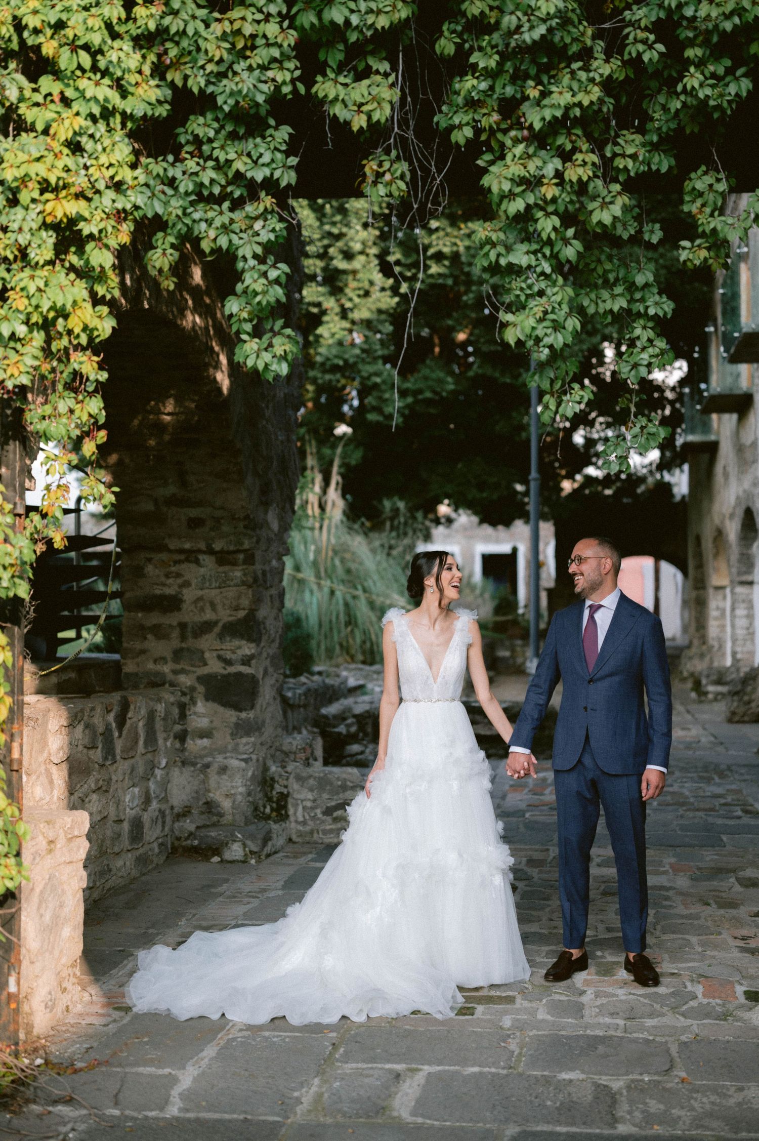 Bride and groom walk hand in hand along ivy-covered stone pathway in charming rustic outdoor setting.