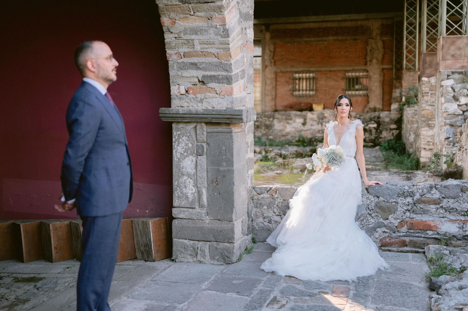 Groom in navy suit watches bride in layered tulle wedding gown at historic ruins venue.