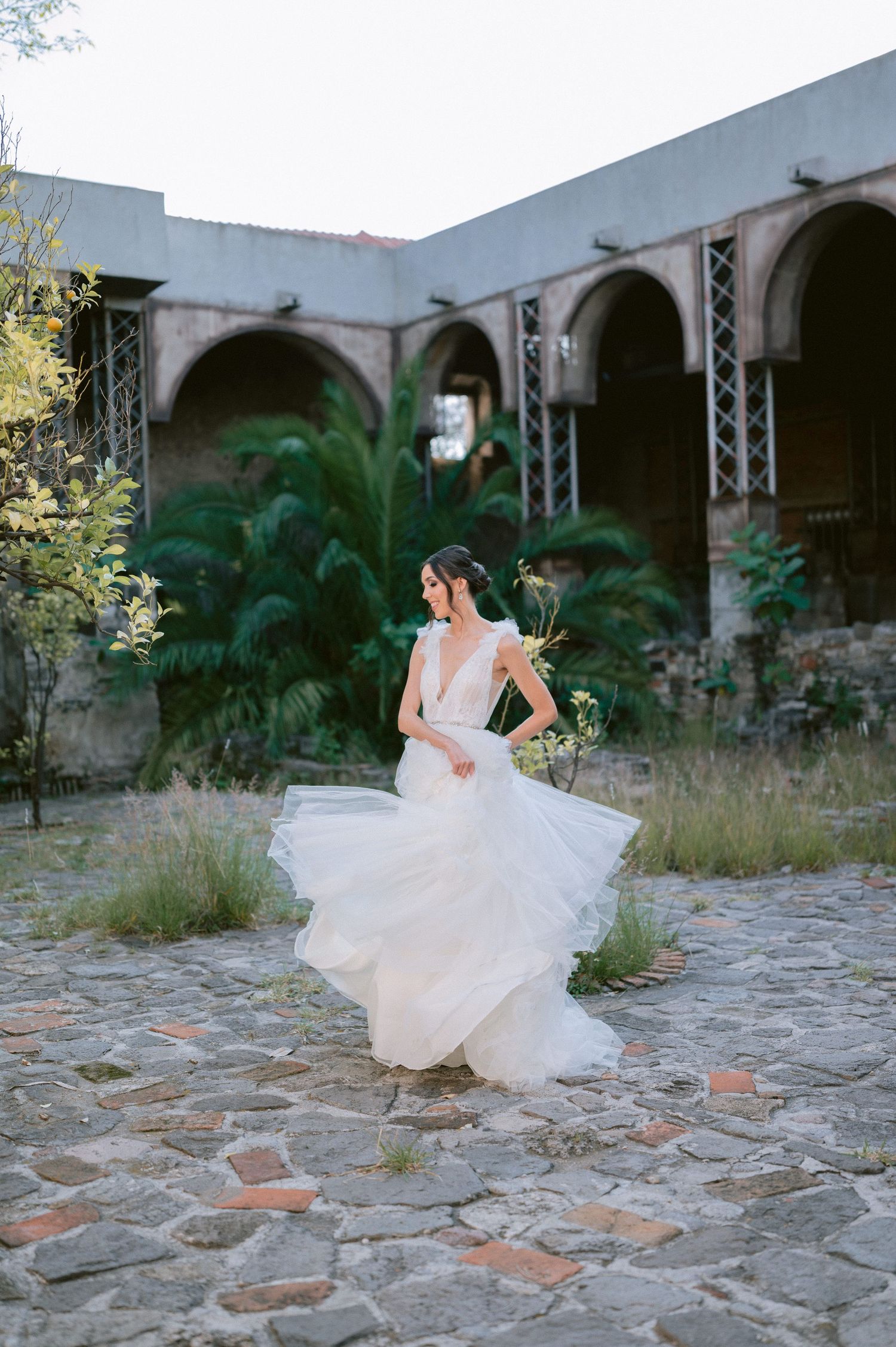Bride in elegant tiered tulle wedding dress twirls on stone courtyard surrounded by arched historic building.