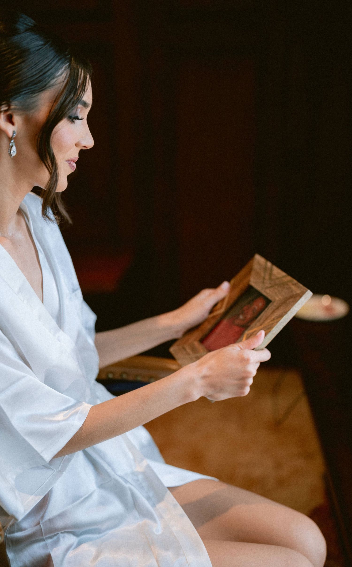 Bride in white satin robe reading from a framed religious icon during wedding preparations.