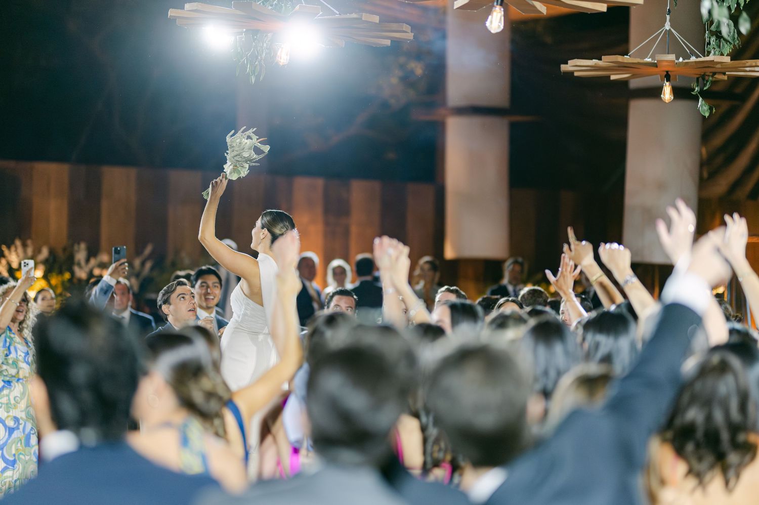 Bride in white gown raises bouquet above crowd of cheering guests during lively indoor wedding reception.