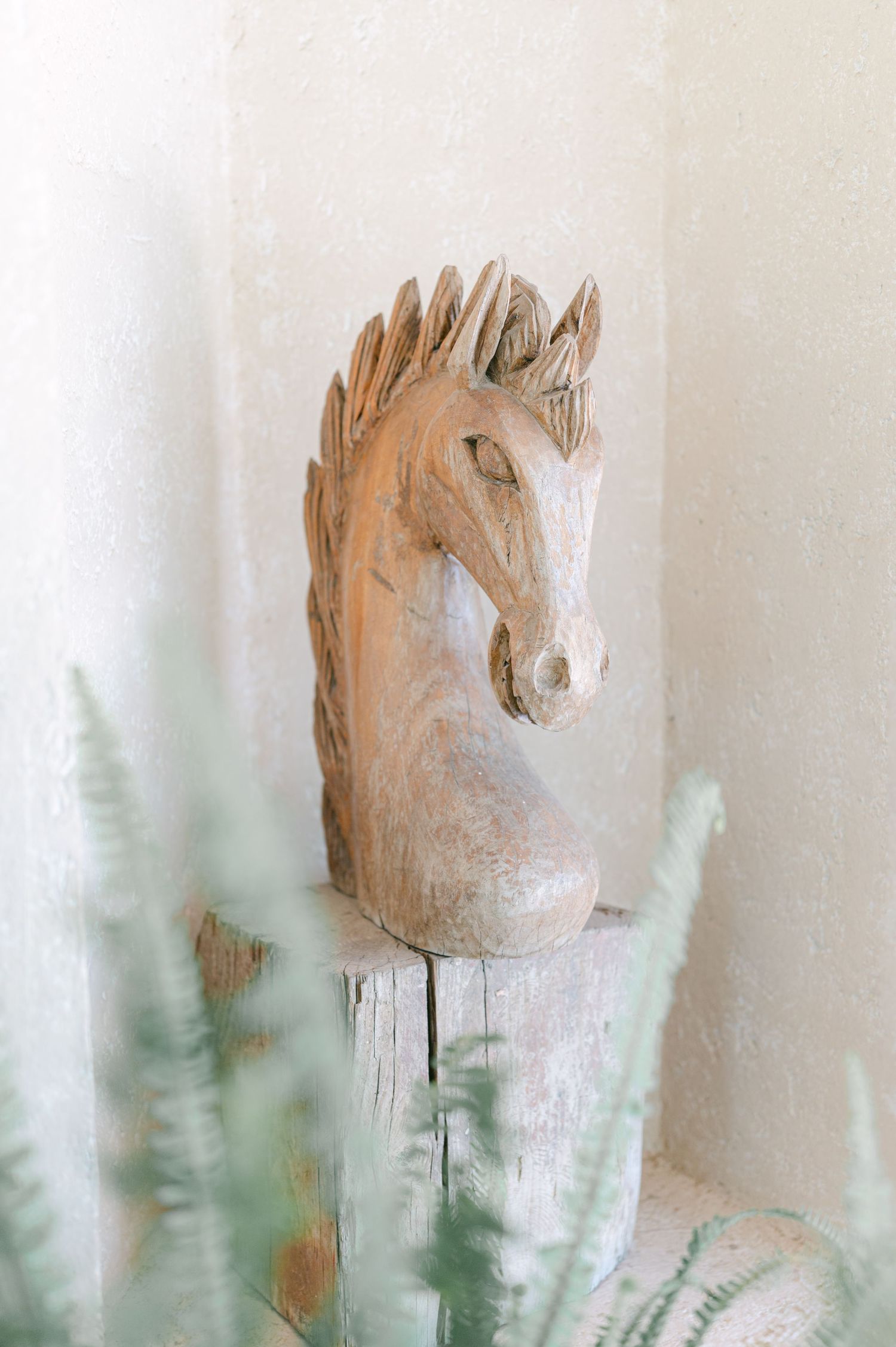 Rustic wooden carved horse head sculpture displayed on a pedestal with soft green foliage in foreground.