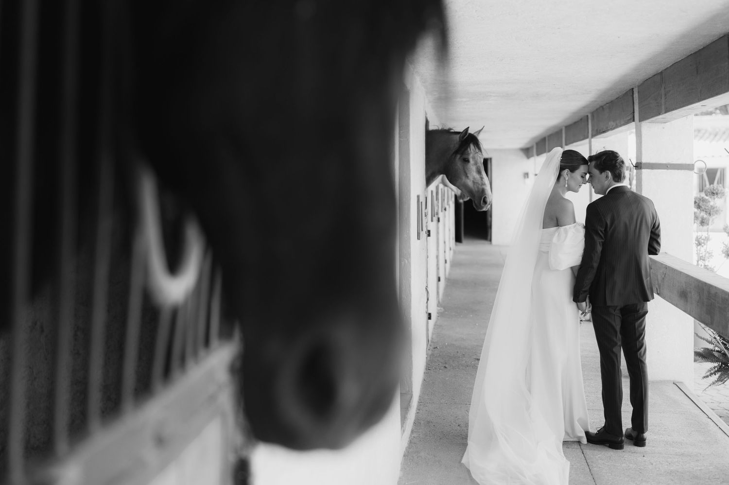 Black and white photo of couple sharing moment near horse stables with horse peering out.