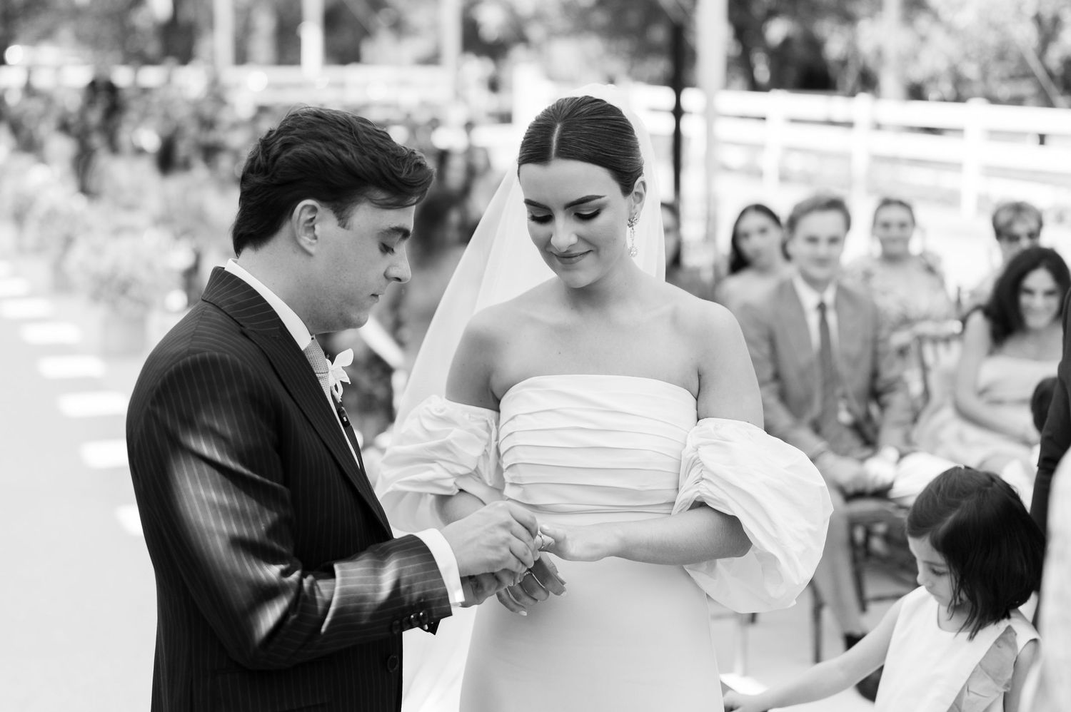 Black and white photo of a groom placing a ring on his bride's finger during an outdoor wedding ceremony.