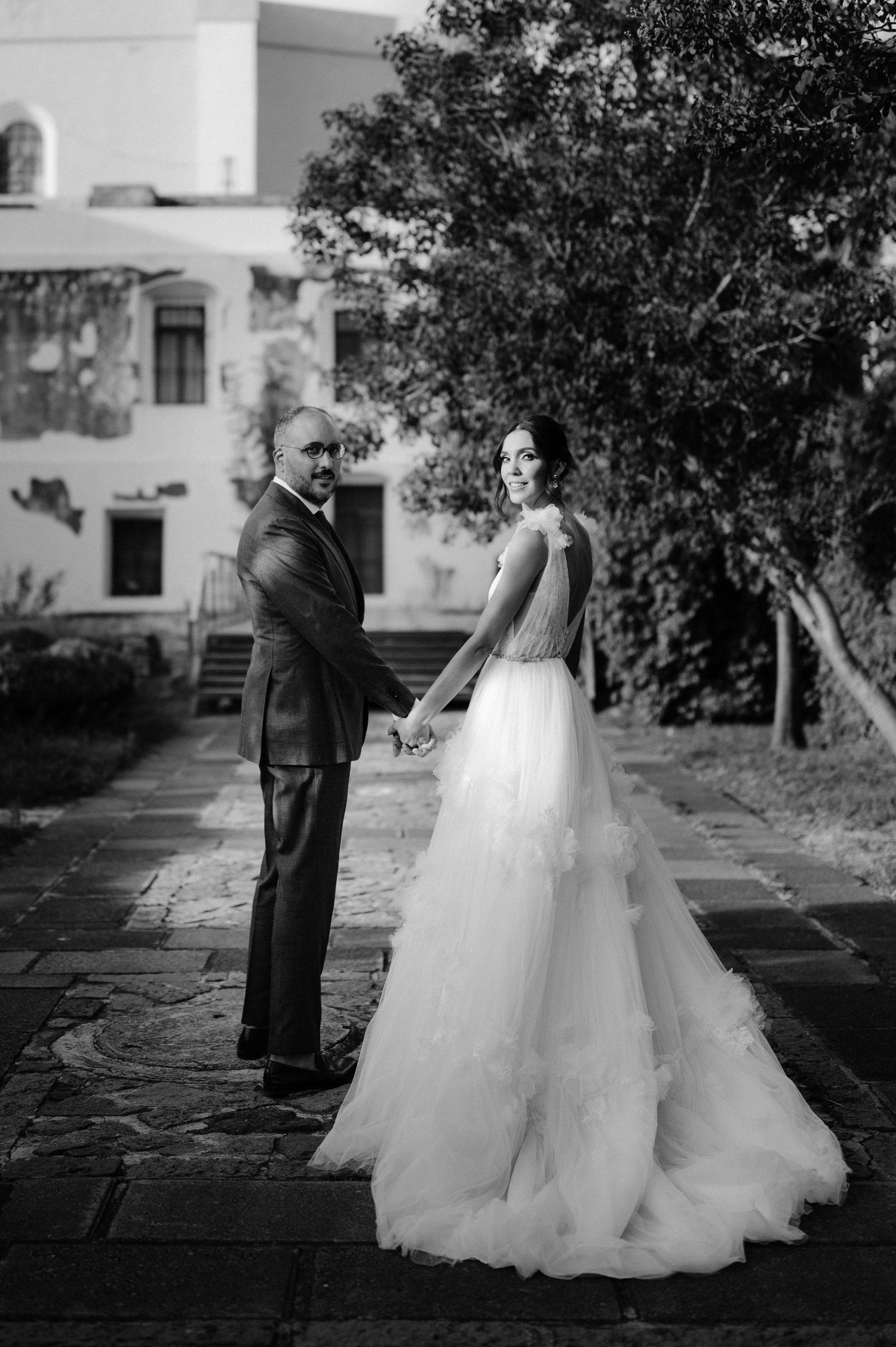 Black and white photo of bride in flowing gown and groom holding hands at outdoor venue.