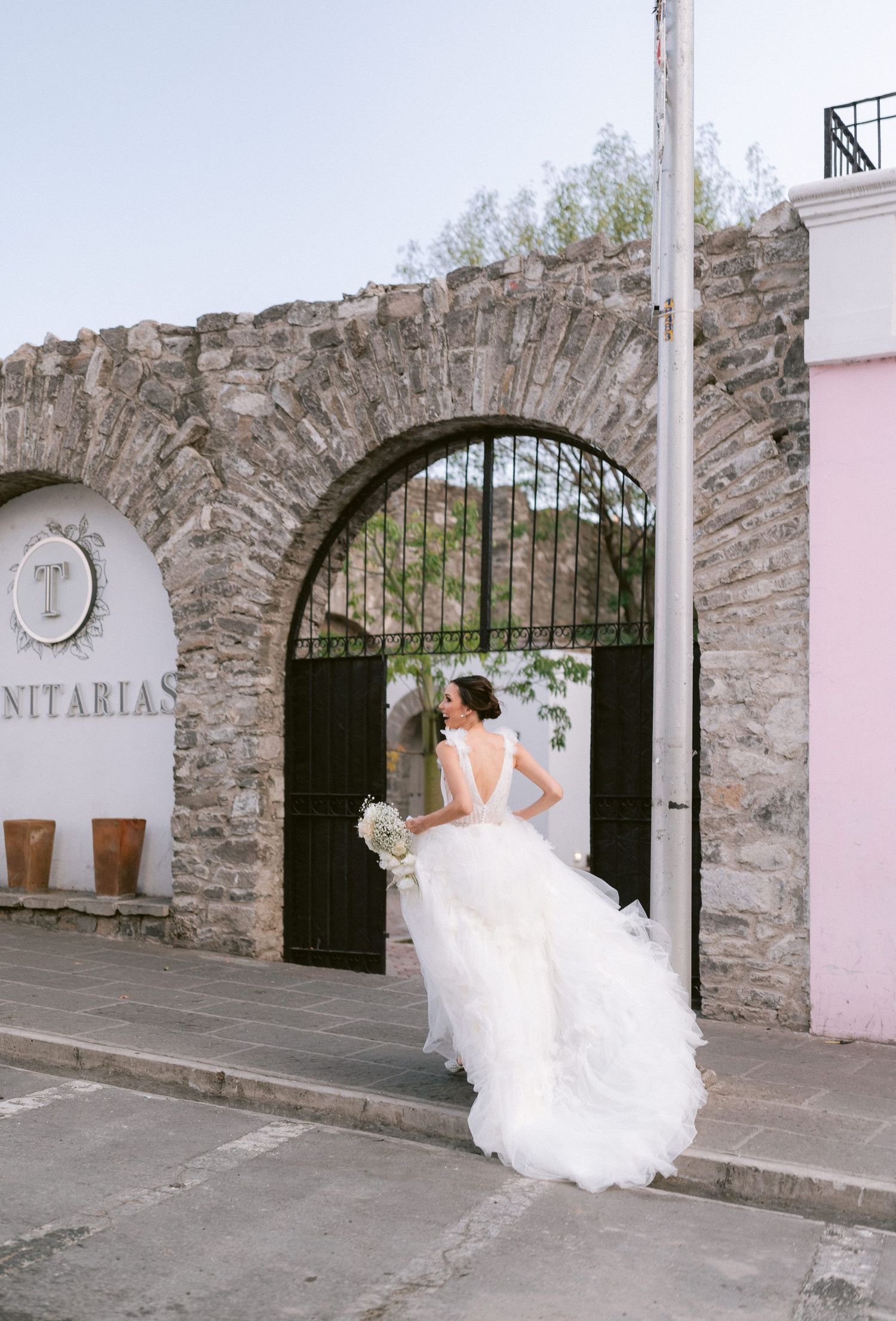 Bride in layered white wedding dress with long train at Trinitarias stone arch gate entrance.