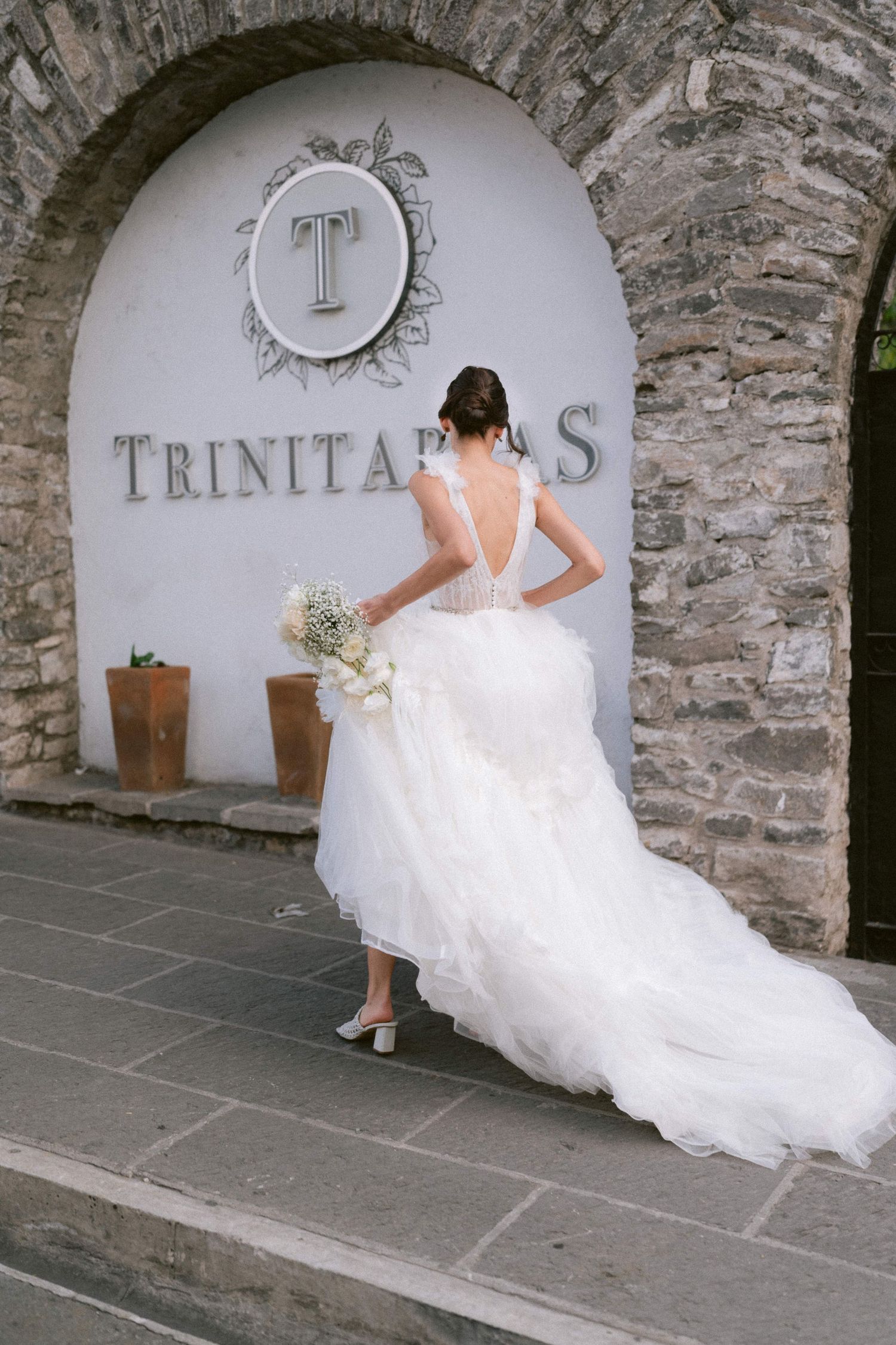 Bride in backless white gown holding bouquet standing at Trinitarias venue stone arch entrance.