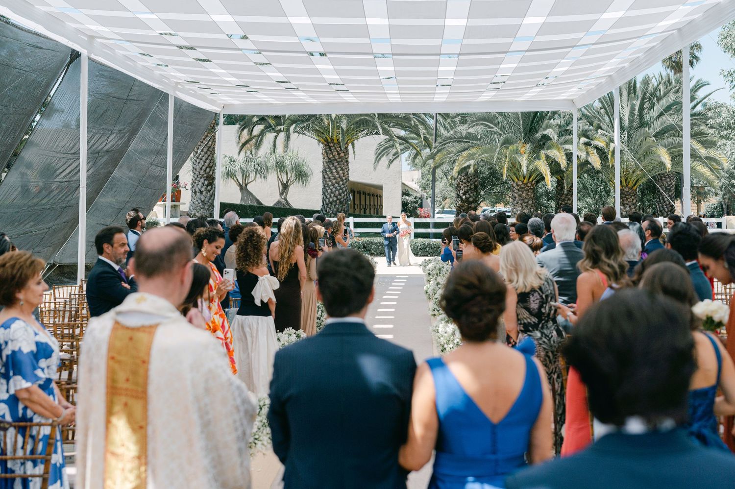 Guests watch bride and groom at altar during outdoor wedding ceremony under white canopy.