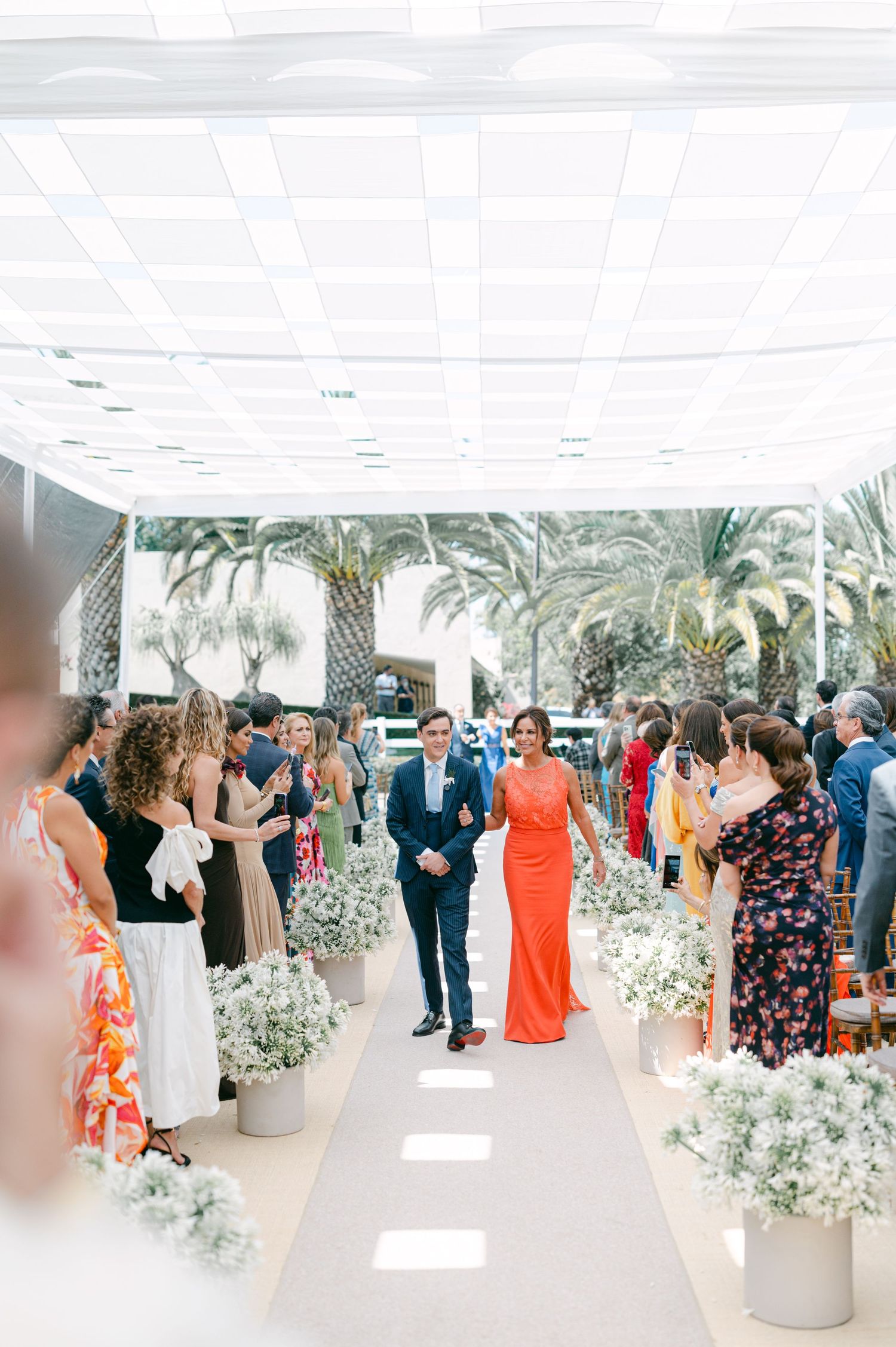 Groom in blue suit walks down aisle with woman in orange dress at elegant outdoor wedding.