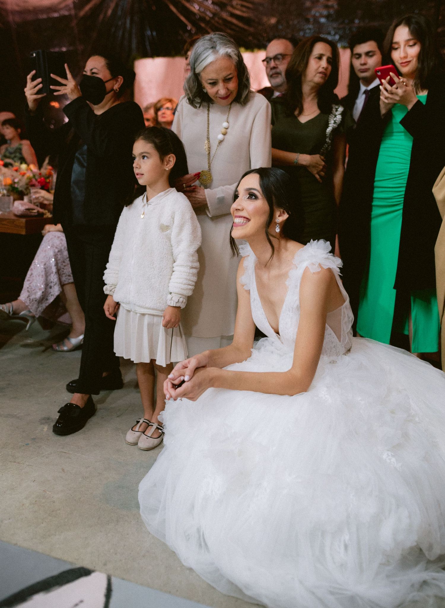Bride in white tulle gown crouches beside young flower girl while wedding guests gather around them.
